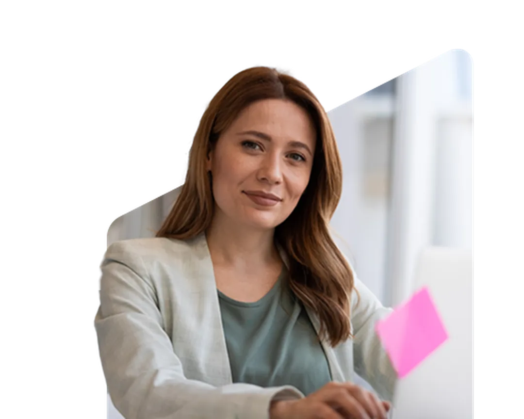 Woman with long hair in a light blazer and green top, sitting at a desk with a laptop and a pink sticky note, smiling slightly.