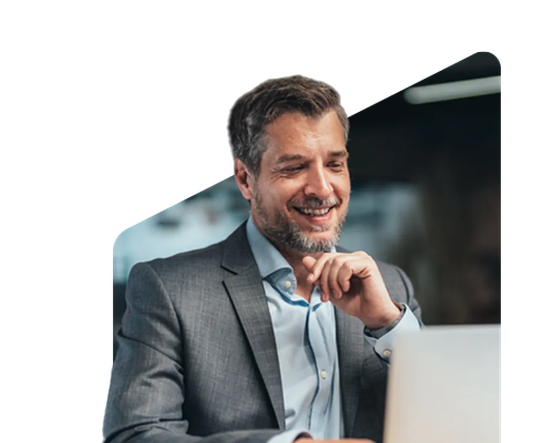 Smiling man in a suit looks at a laptop screen, sitting at a desk in a dimly lit room.
