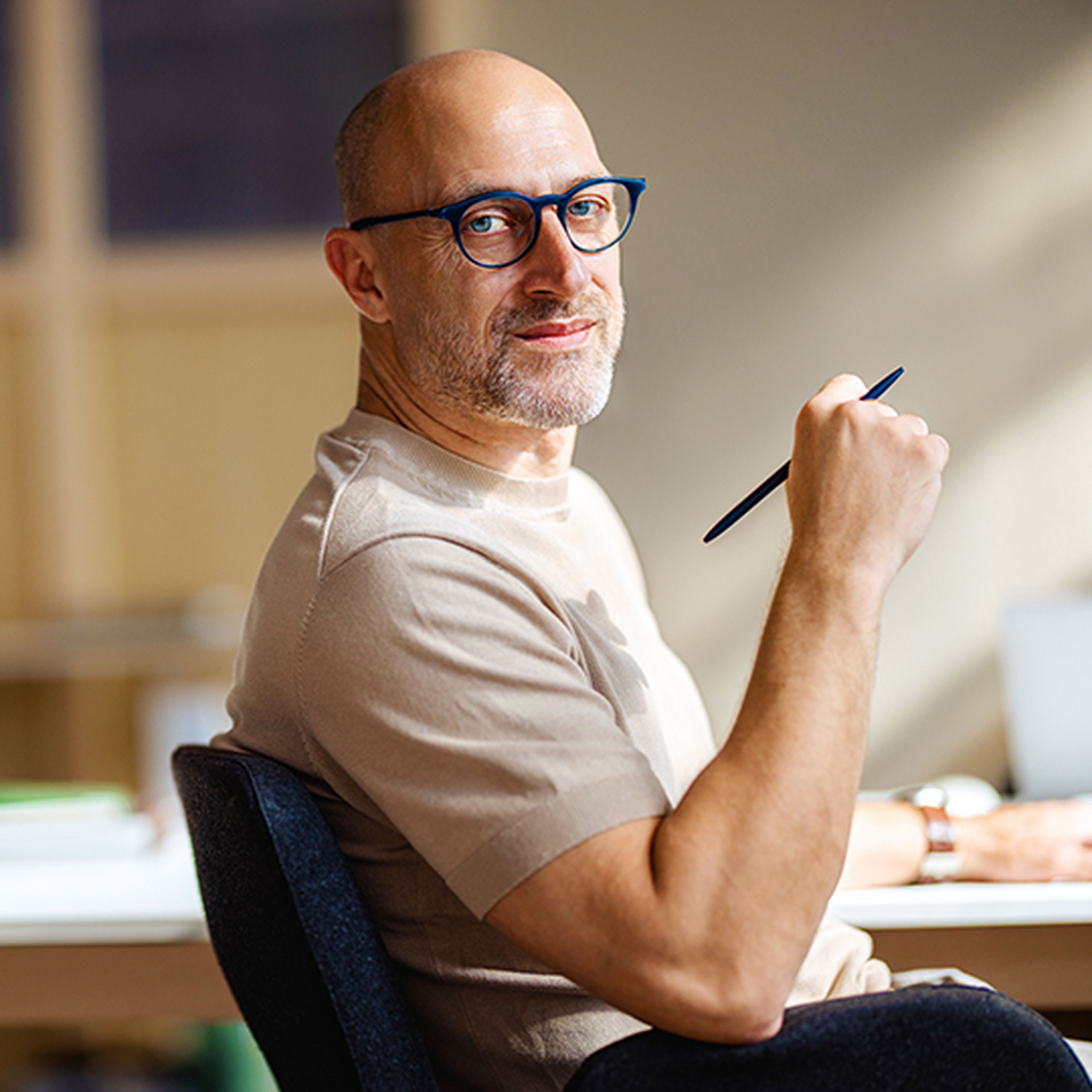 Man with glasses holding a pen, seated at a desk, looking back and smiling. Bright, sunlit room with blurred background.