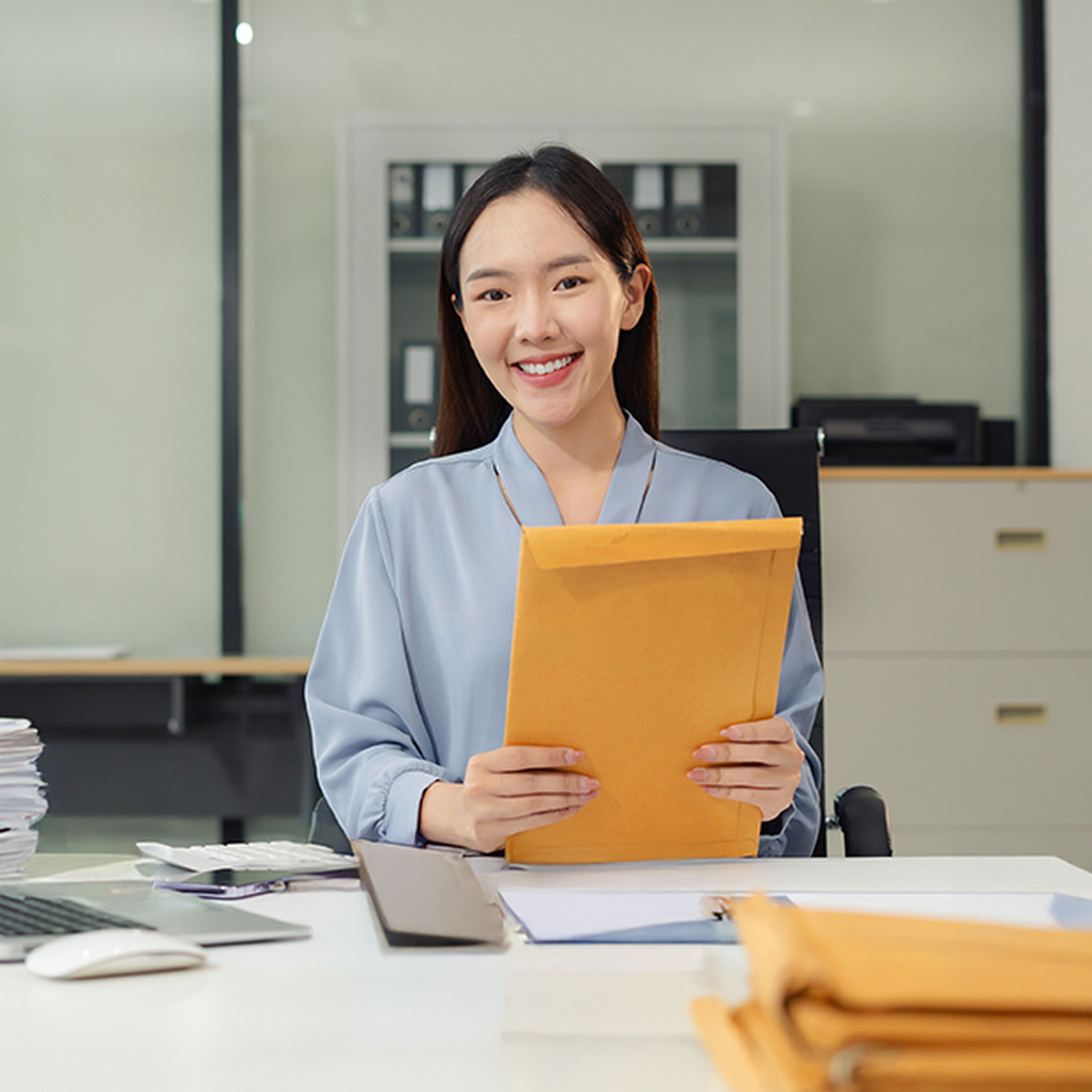 Smiling woman in a blue blouse holding a yellow envelope, sitting at a desk with papers and folders in an office setting.