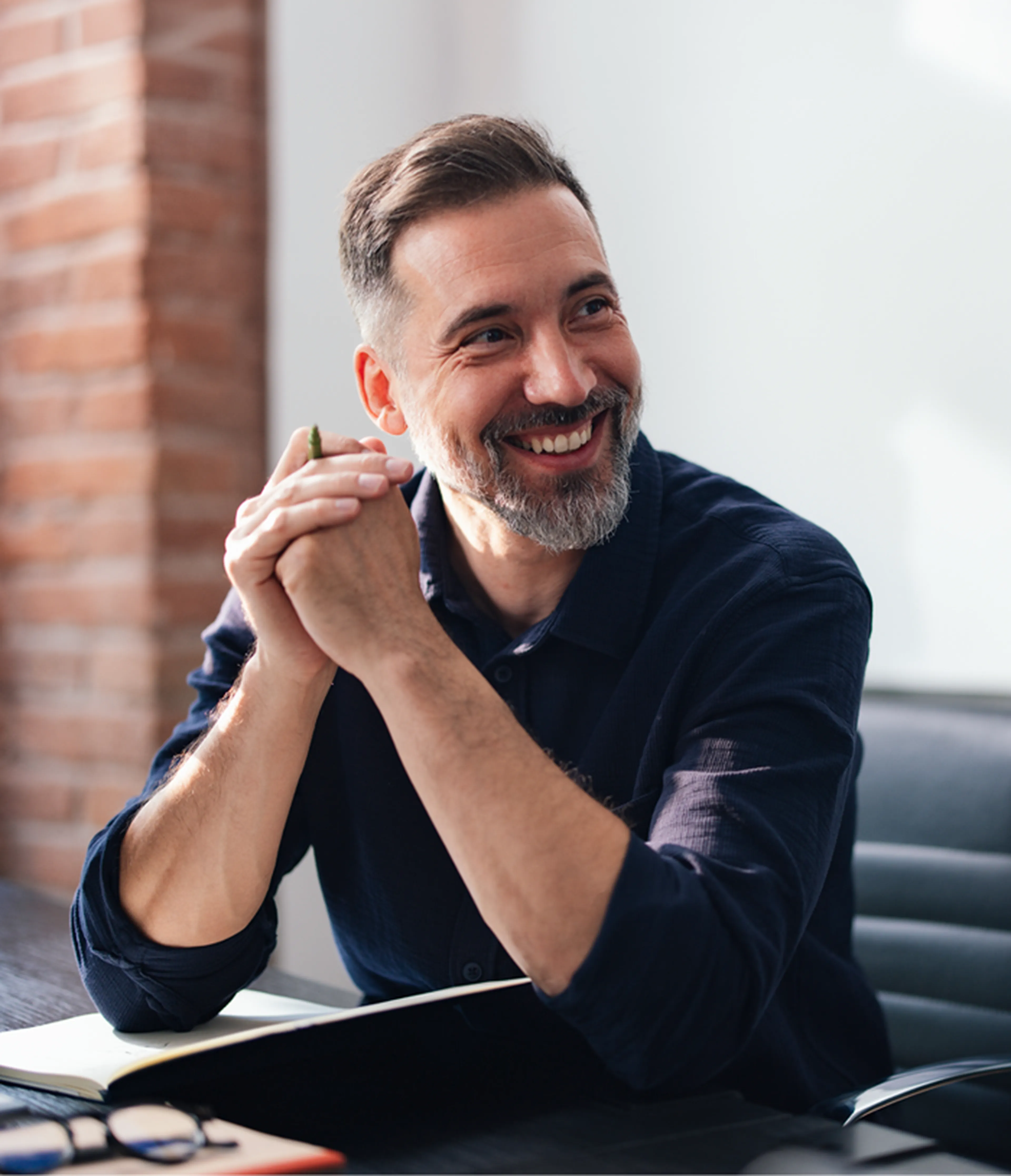 Smiling man with a beard sitting at a desk, hands clasped, next to an open notebook and glasses, in a well-lit room with a brick wall background.