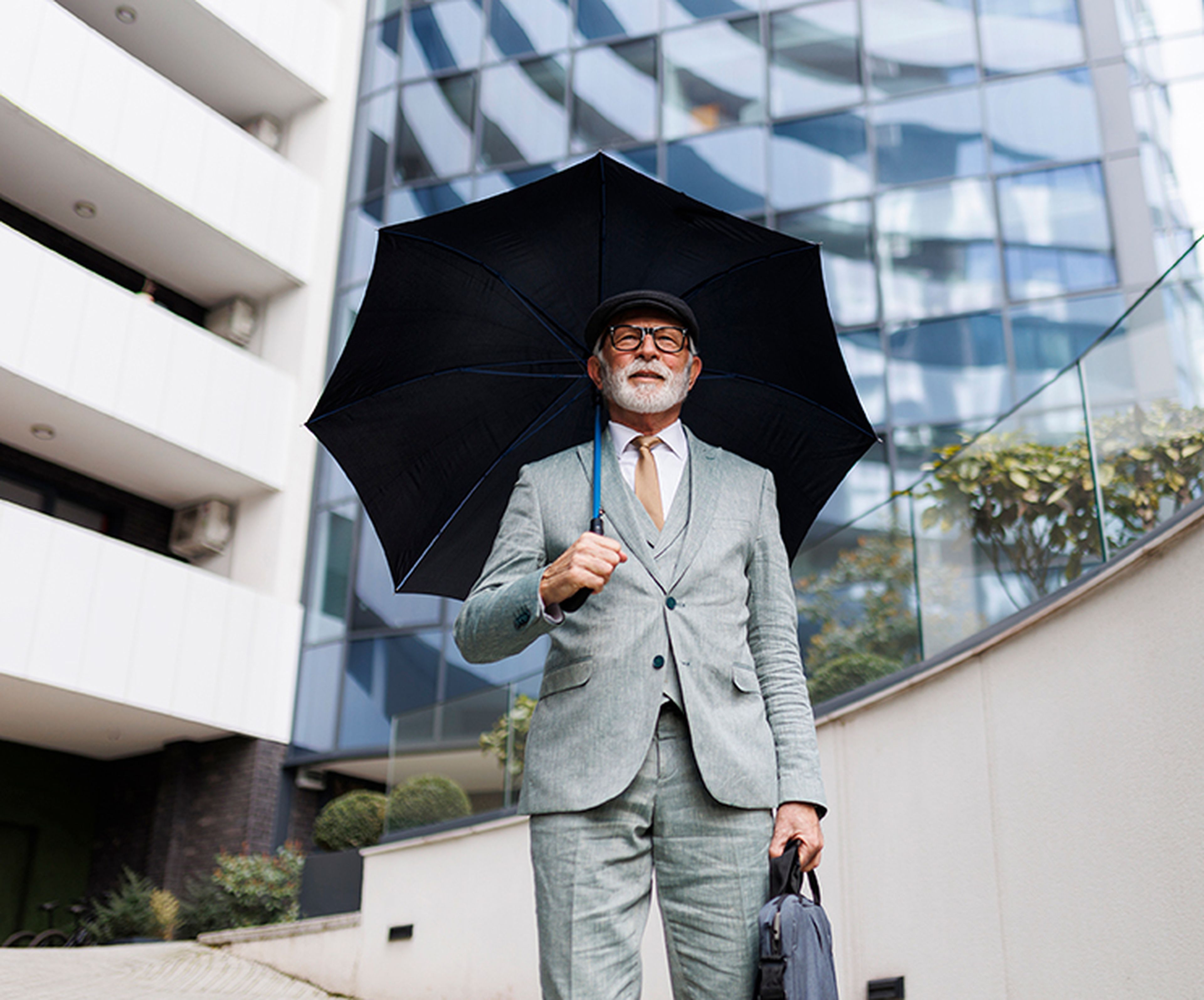Experienced man in a light gray suit holds a black umbrella, standing in front of modern glass buildings.