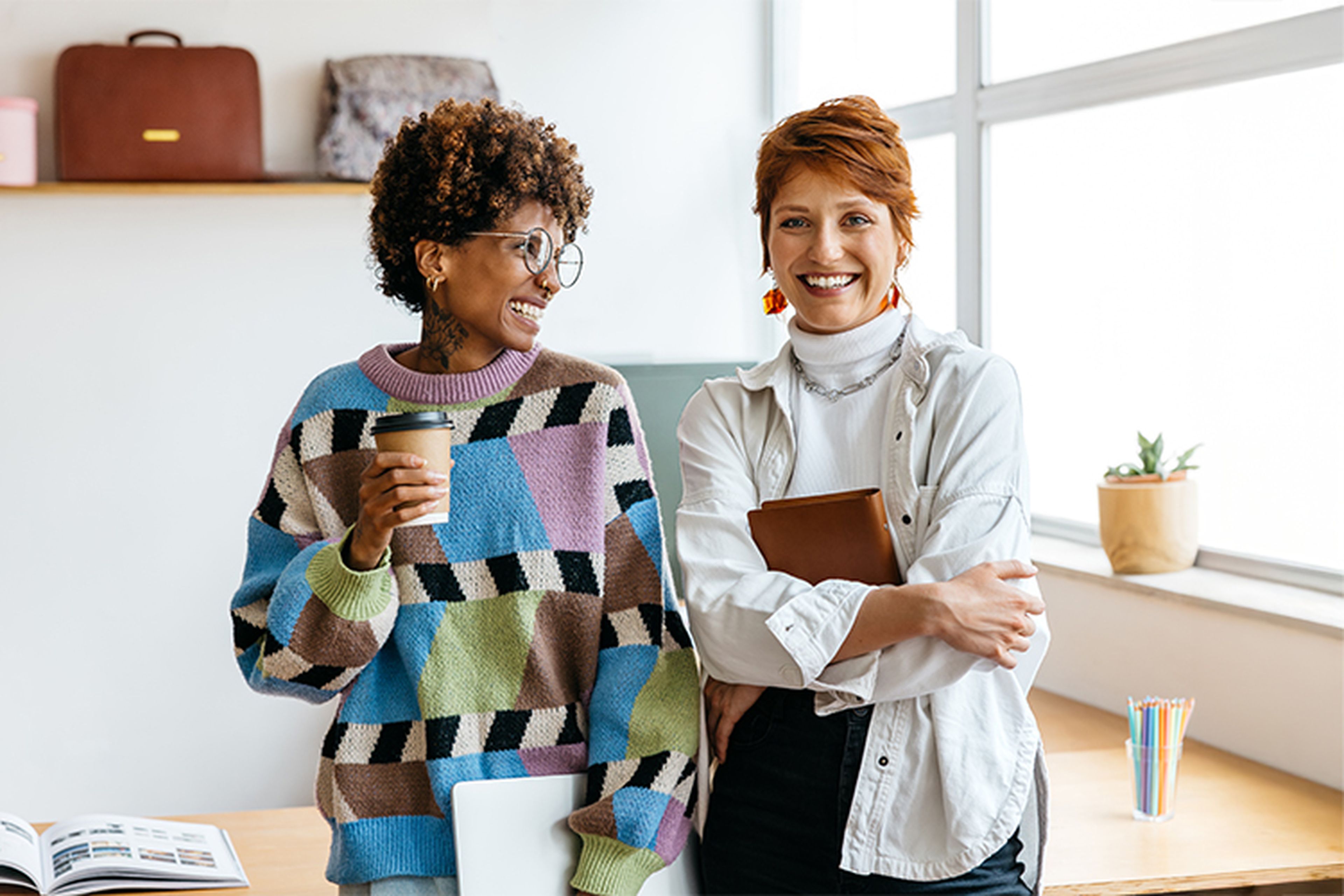 Two people smiling in a bright office. One holds a coffee cup, wearing a colorful sweater; the other holds a notebook, wearing a white shirt.