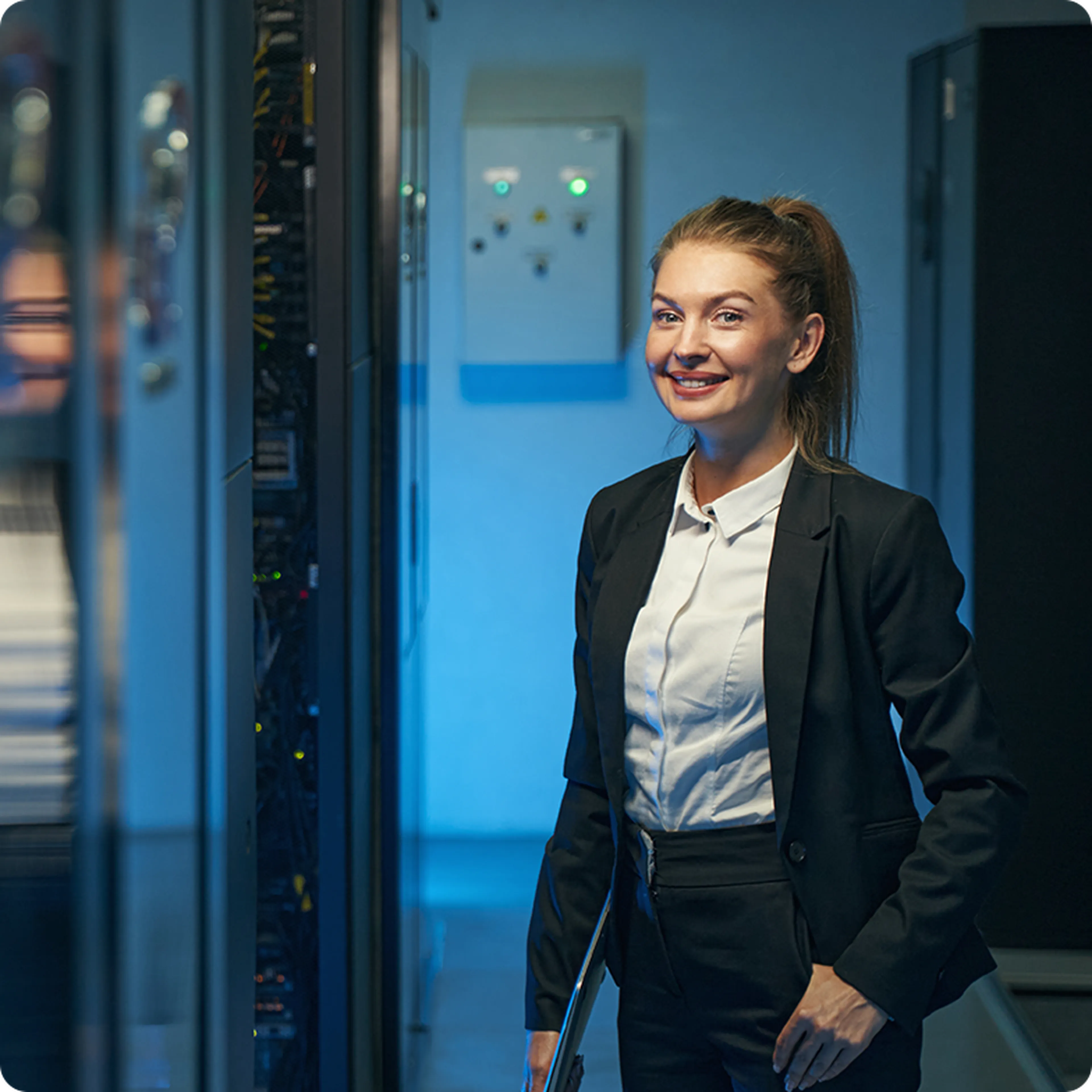 Smiling woman in a suit stands in a server room, surrounded by computer equipment and blue lighting.