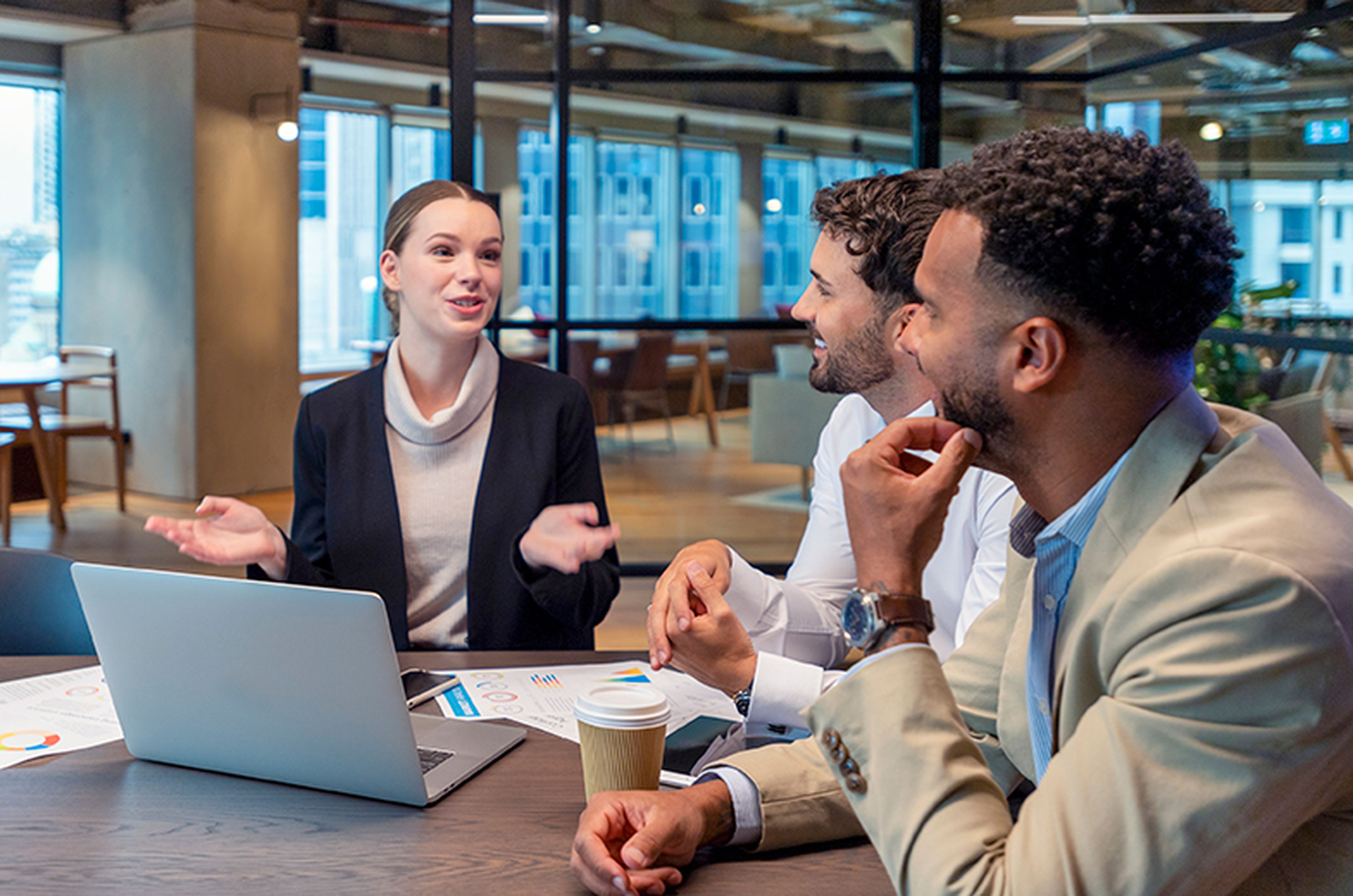Three business people in a modern office discussing work around a table with a laptop, documents, and a coffee cup.