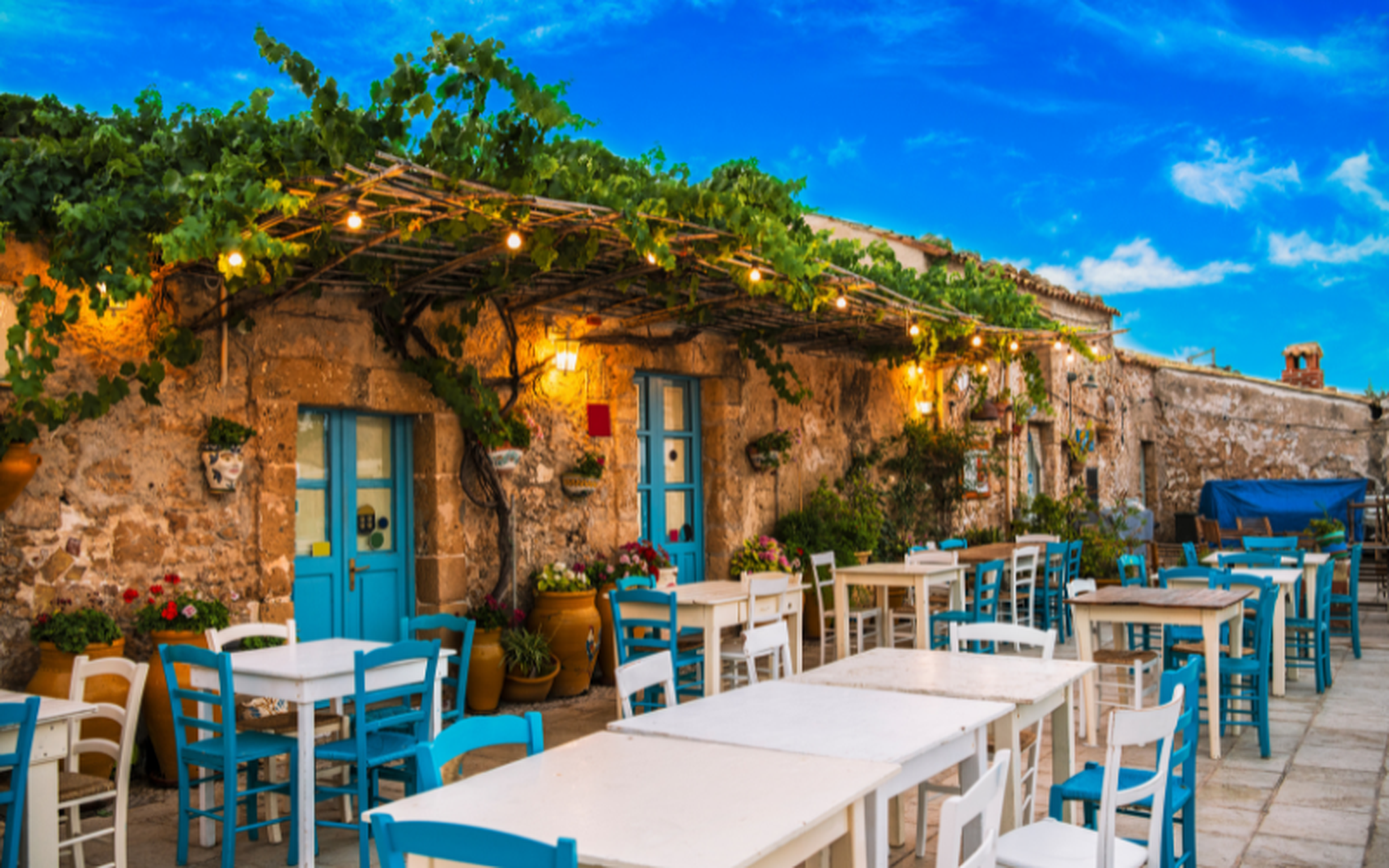 Outdoor café with colorful tables and chairs in a scenic Mediterranean village, framed by traditional stone architecture.