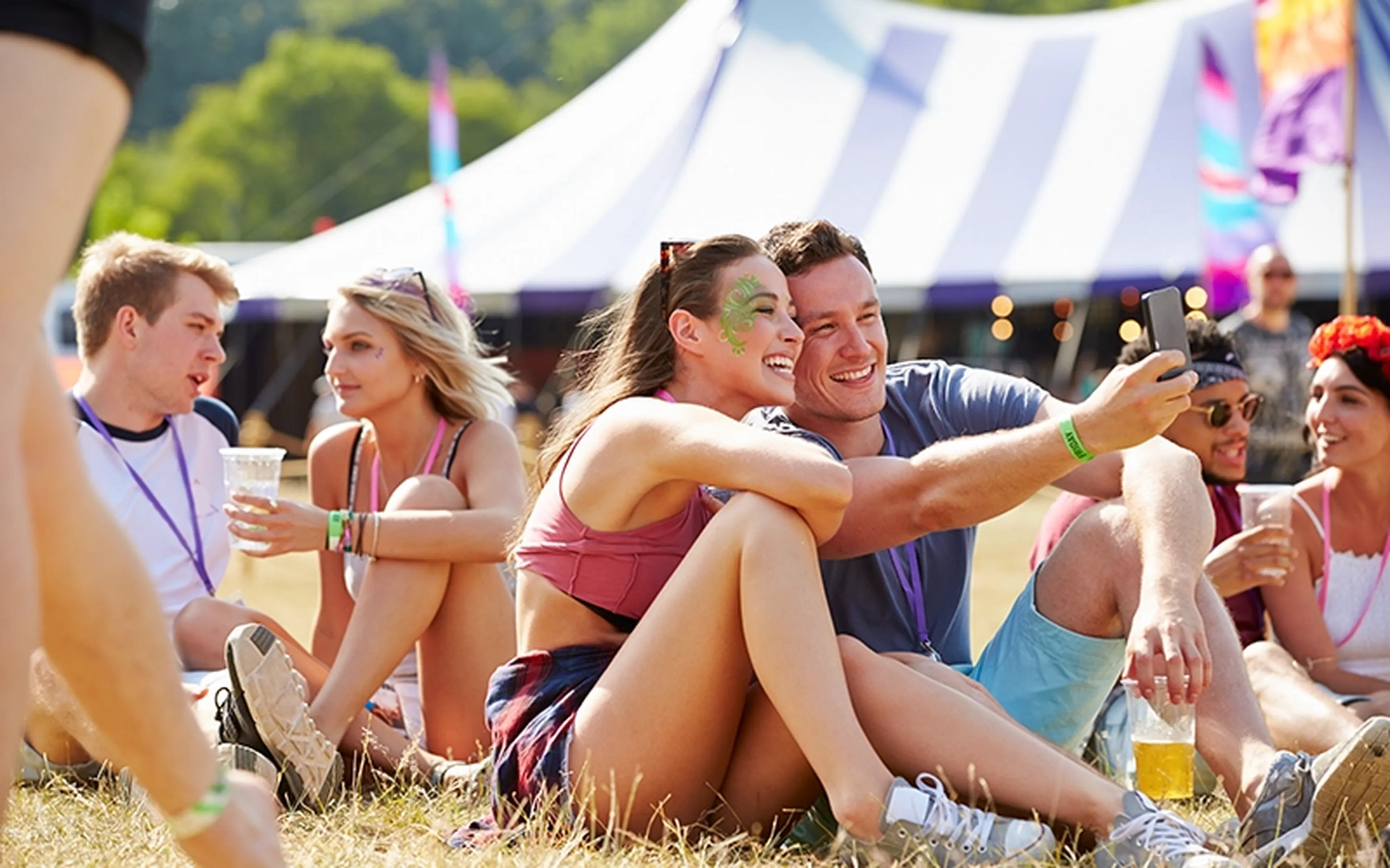 Friends sitting on the grass at an outdoor festival smile while taking a selfie together