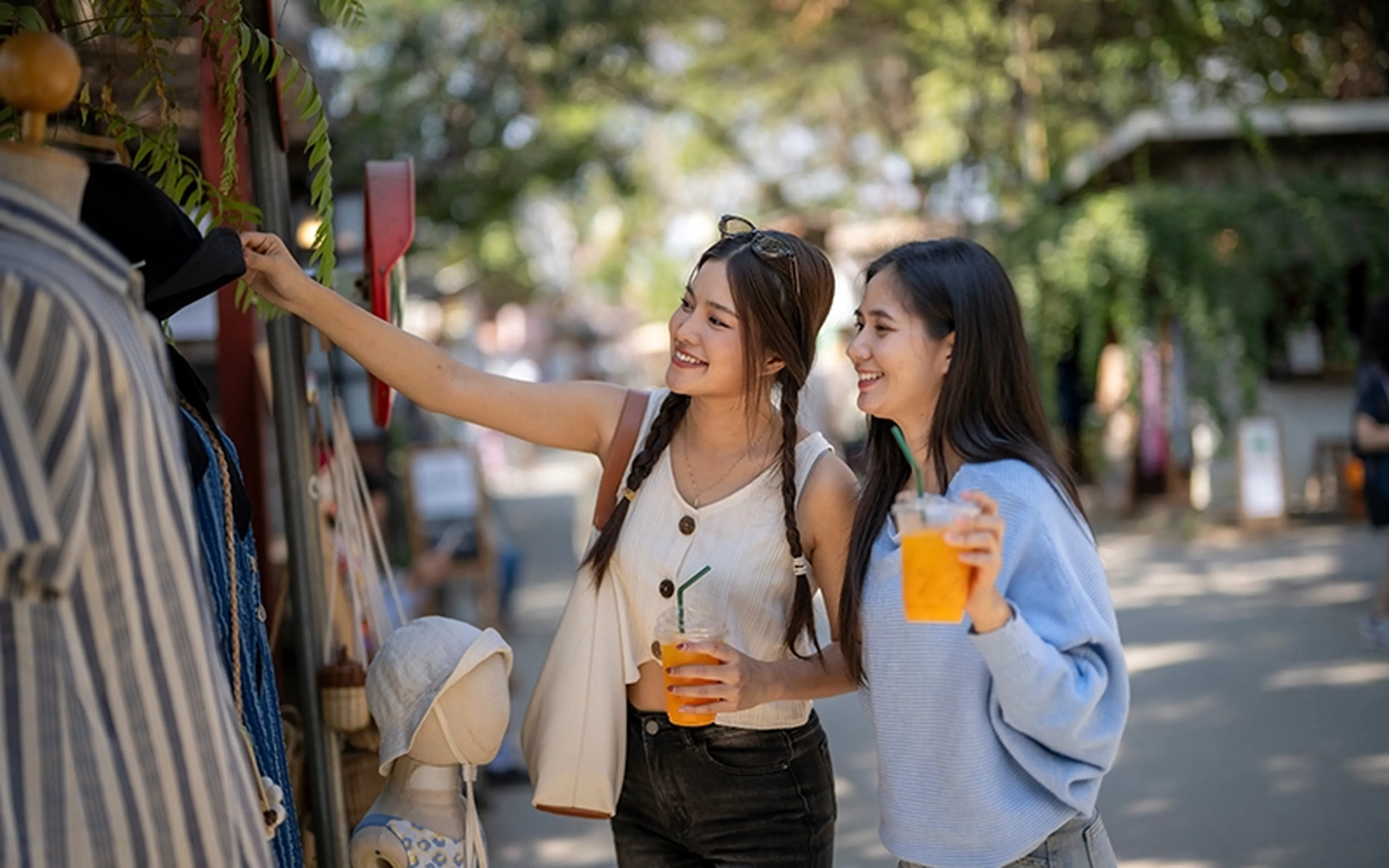 Two friends smile while browsing clothing at a sunlit street stall, each holding a cold drink