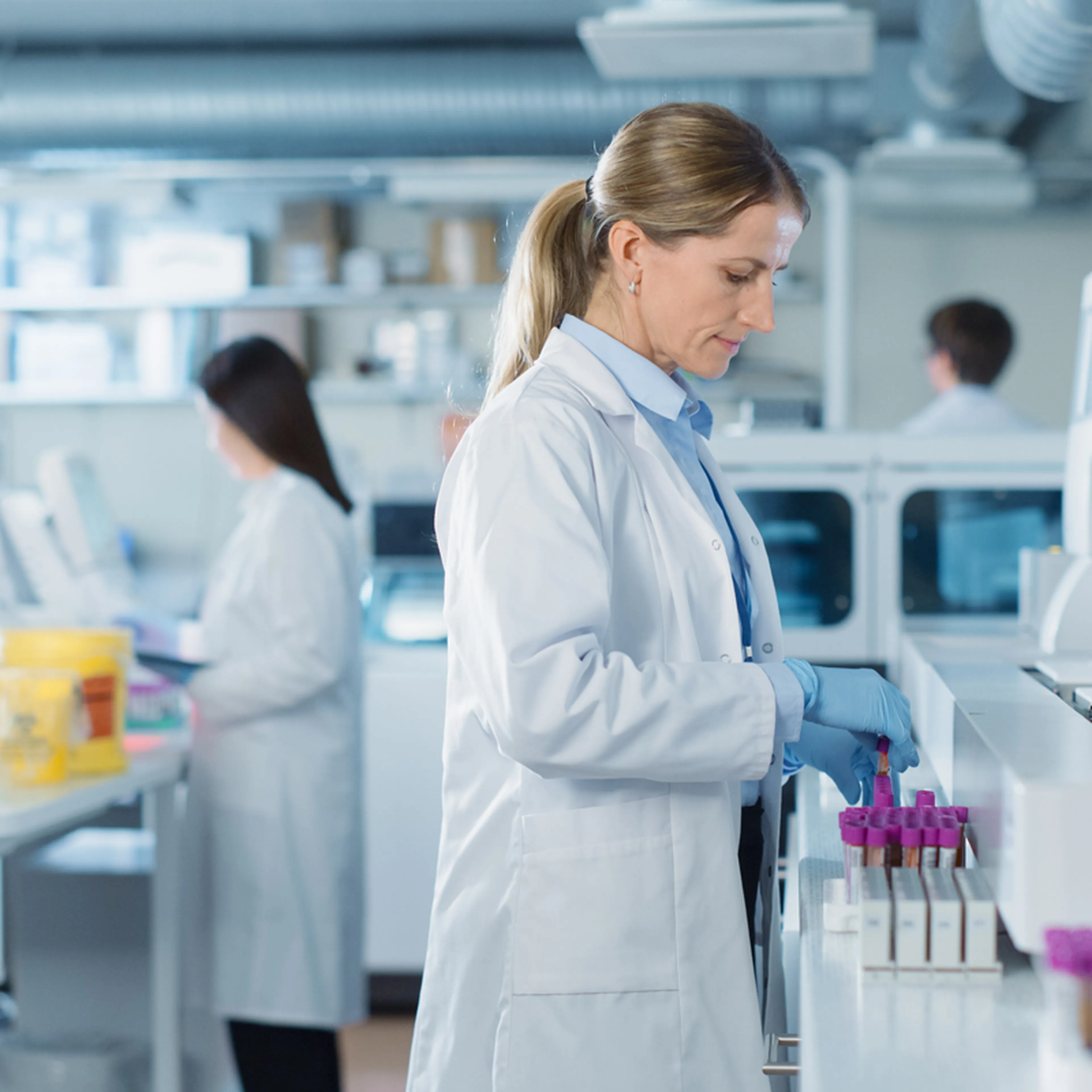 A scientist in a lab coat works with test tubes, while another person in the background operates laboratory equipment.