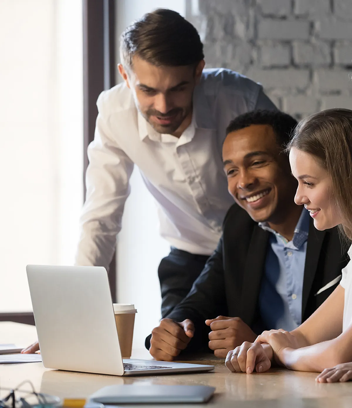 Young team smiling and looking at a laptop in an office setting. A coffee cup is on the table next to them.