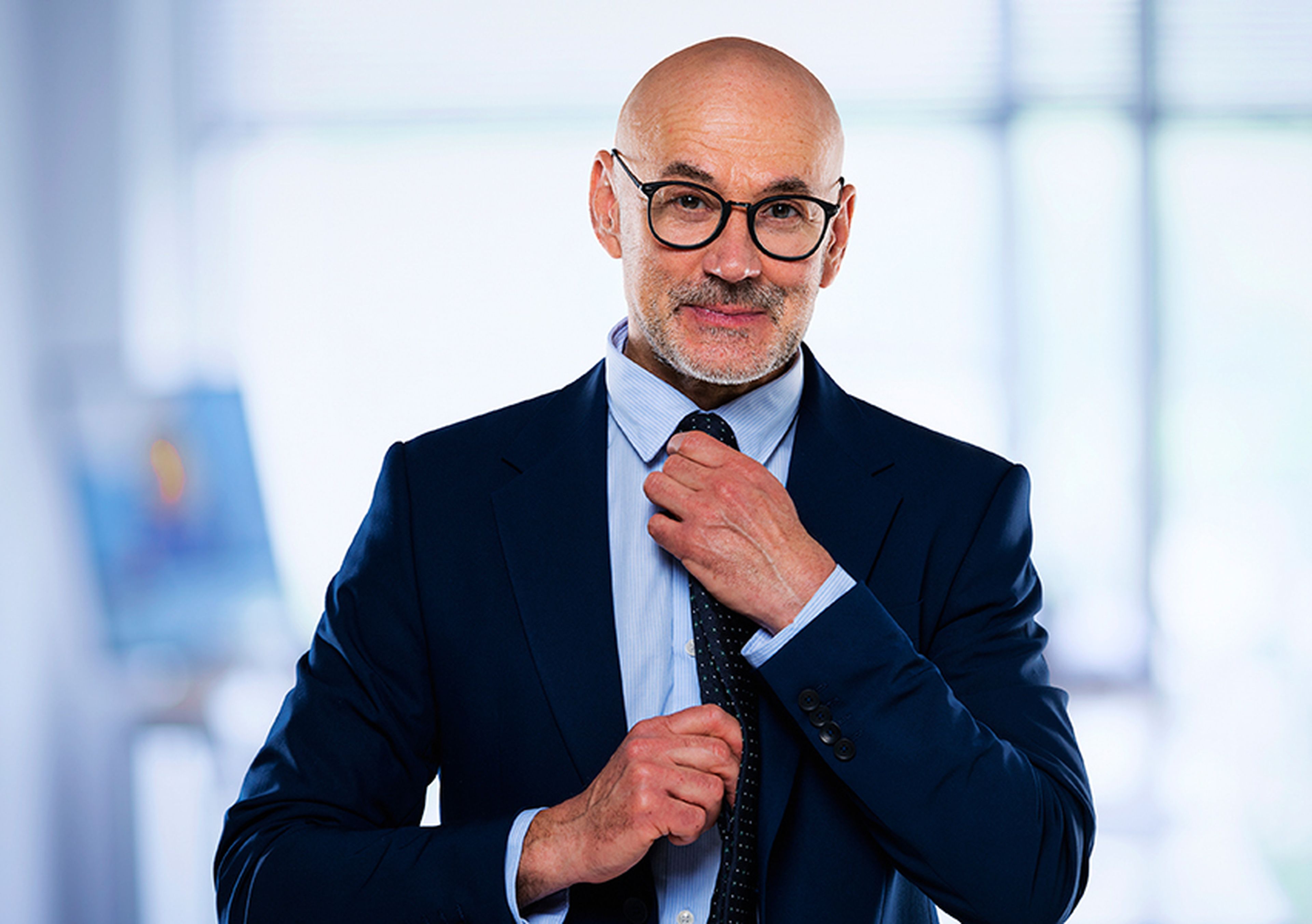 Man with glasses in a suit adjusts his tie, standing in a brightly lit office setting.