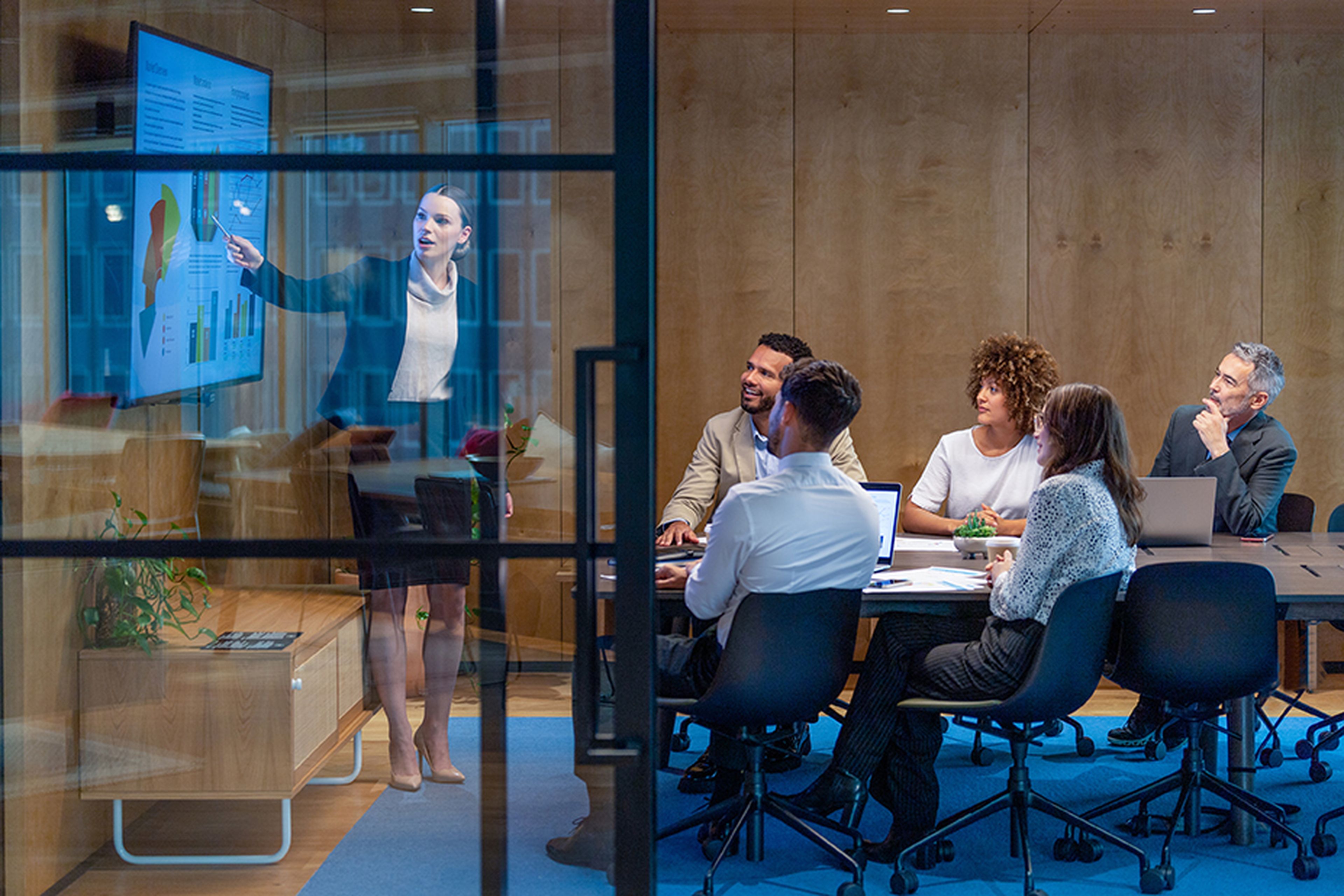 A woman presents data on a screen to five colleagues seated around a table in a modern conference room.