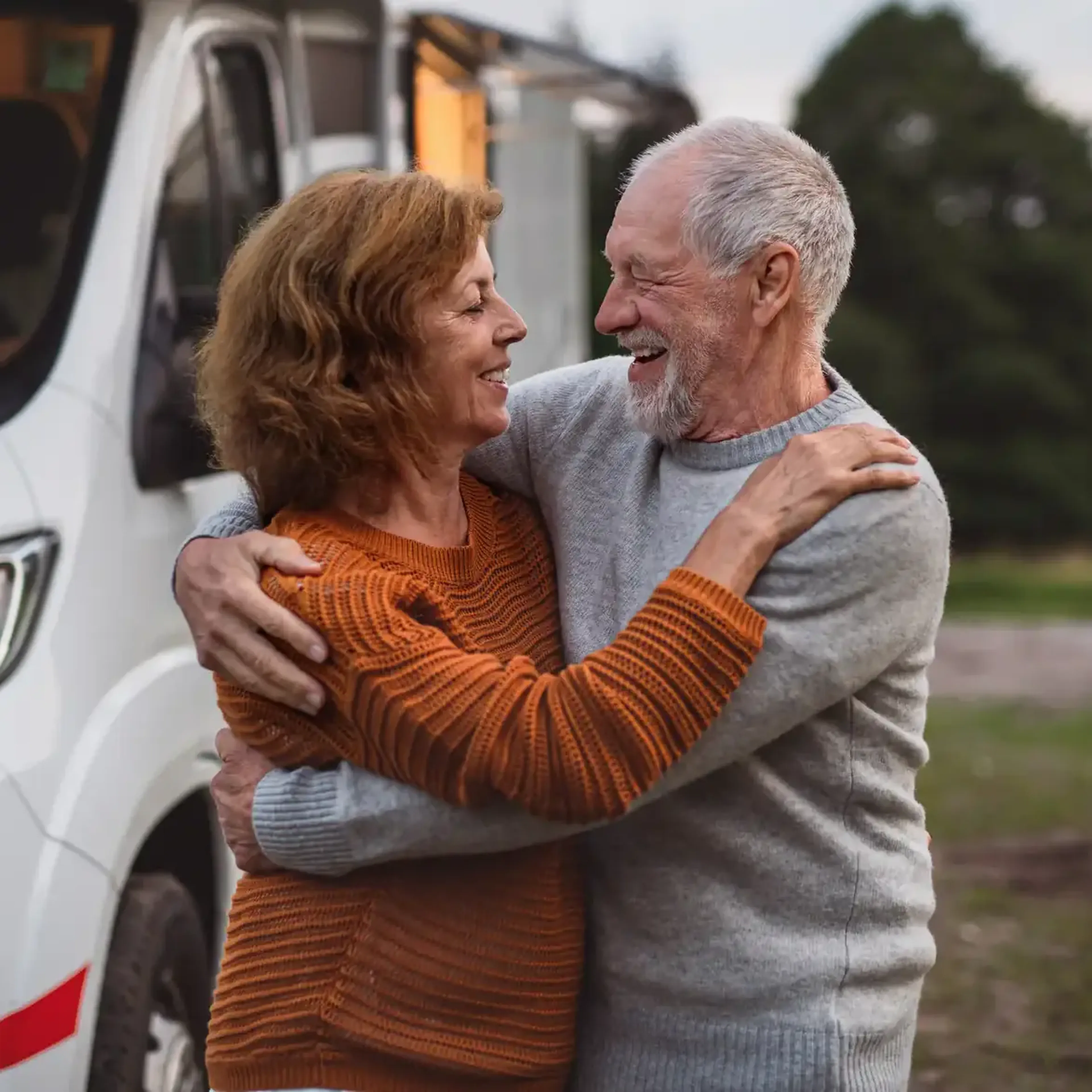 An elderly couple embraces and smiles at each other in front of a camper van, surrounded by trees.