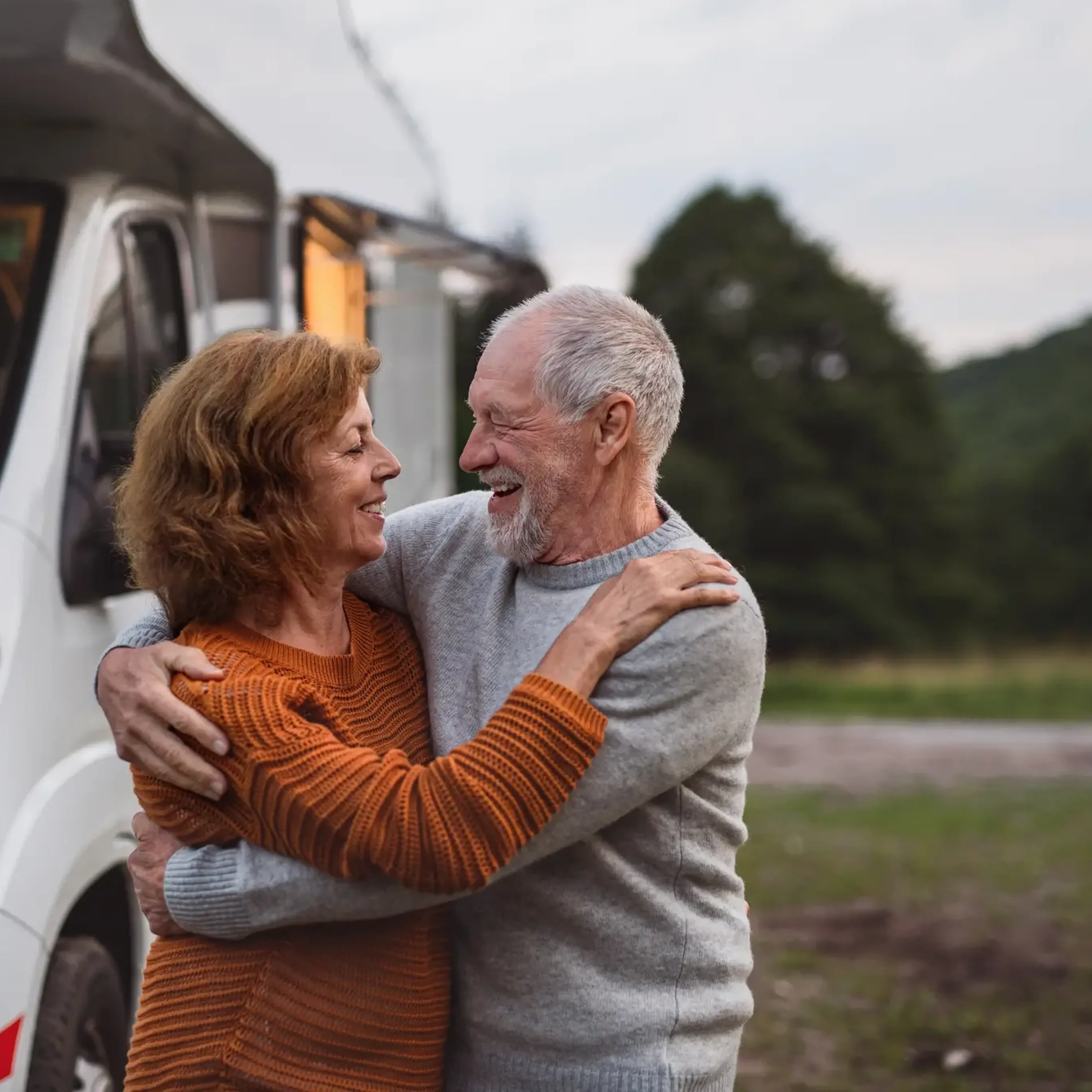 Elderly couple embracing and smiling near a camper van, with trees and hills in the background.