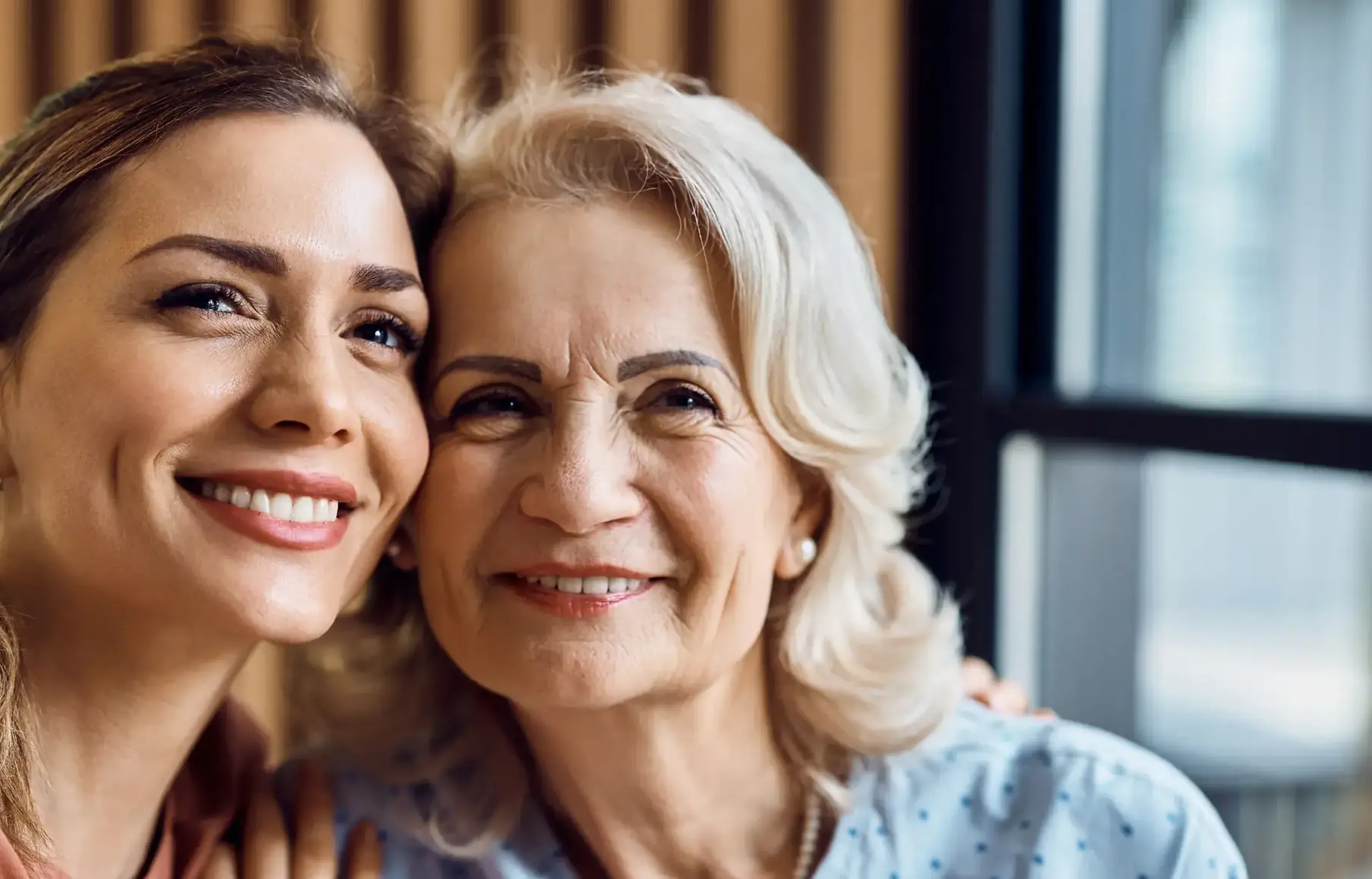 Two women smiling closely together, one younger and one older, in a warmly lit indoor setting.