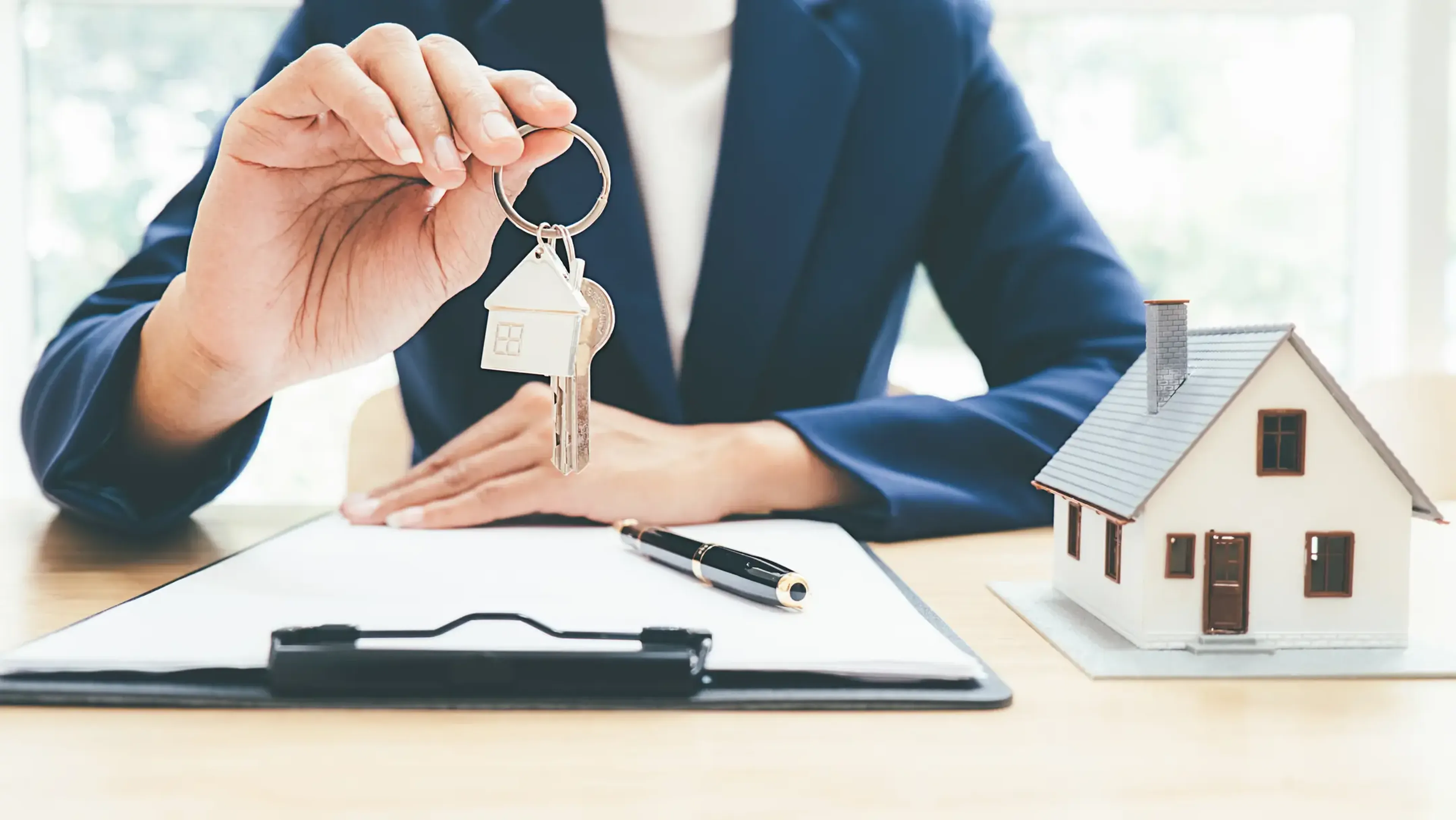 Person in a suit holding house keys over a clipboard, with a pen and a small house model on the table.