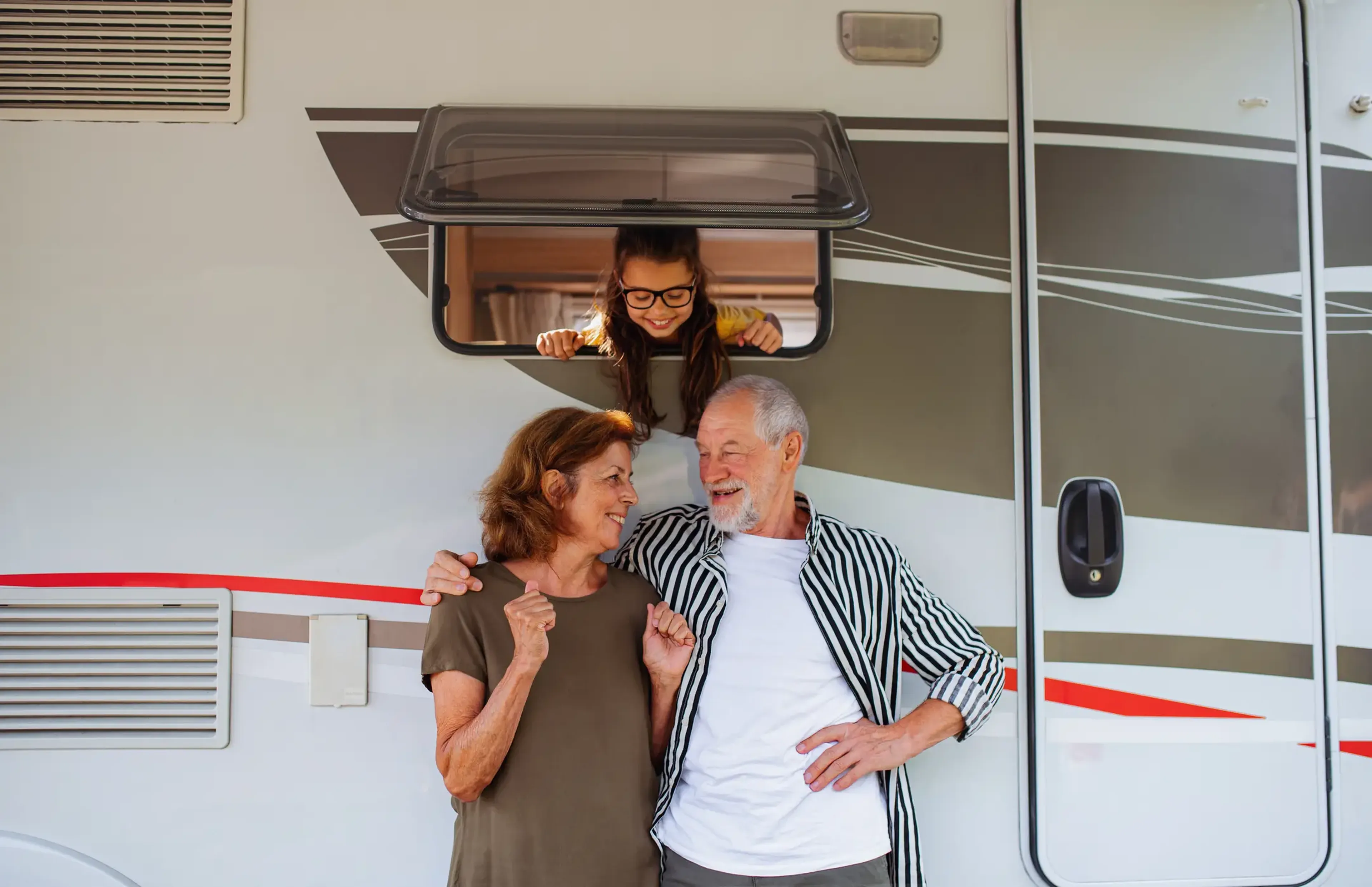 Elderly couple smiling in front of an RV, with a young girl playfully leaning out of a window above them.