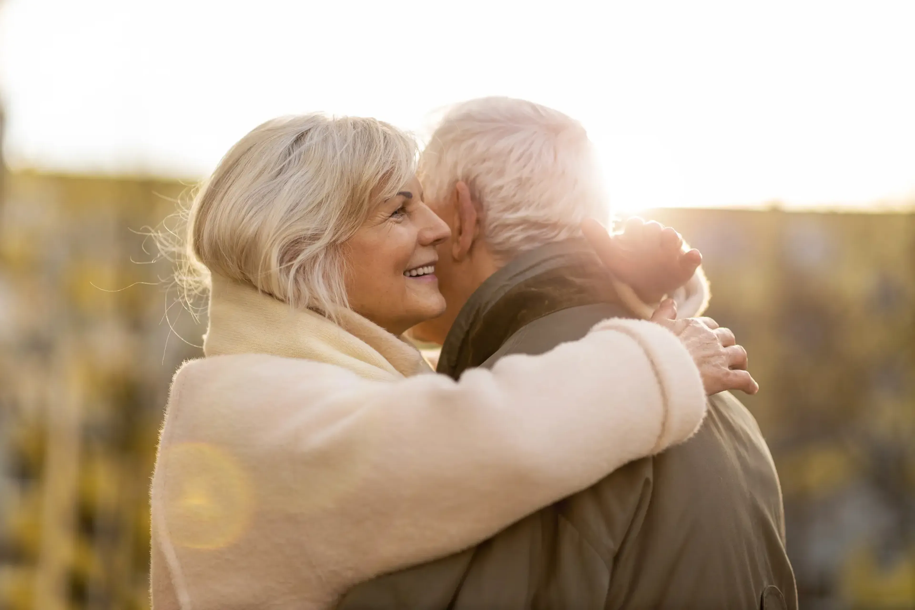 Elderly couple embracing outdoors, woman smiling, with sunlight softly illuminating them from behind.