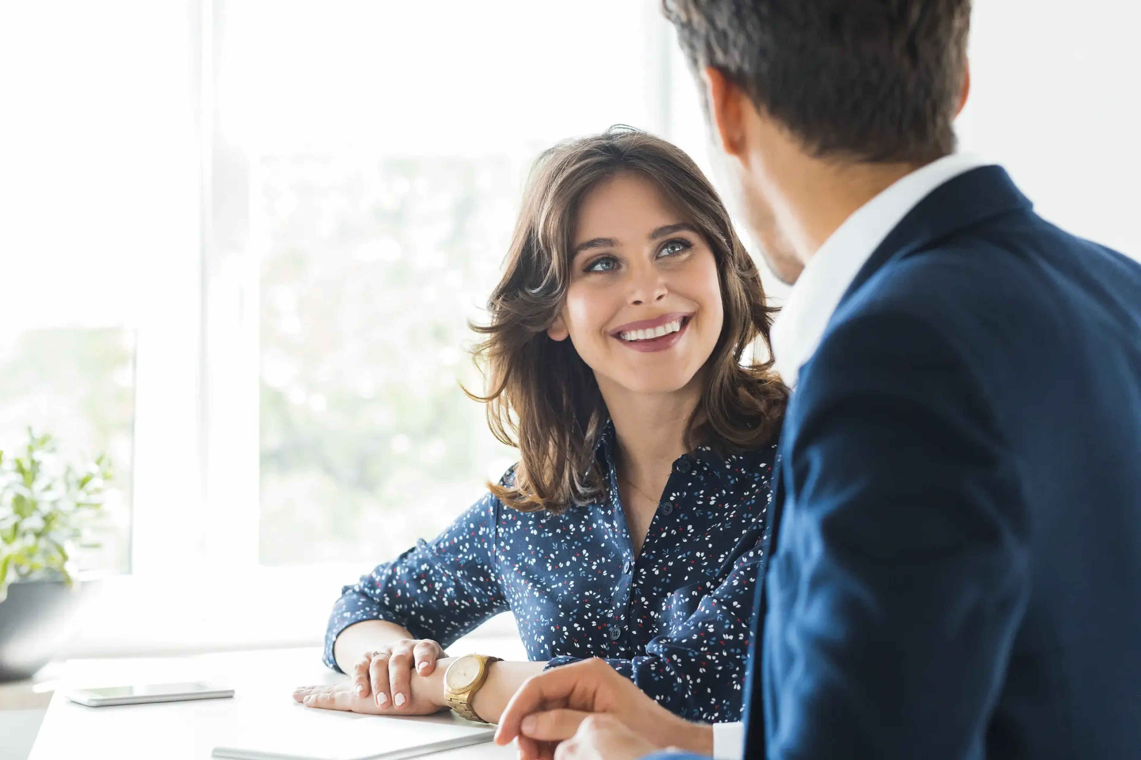 A woman in a blue blouse smiles at a man in a suit during a conversation in a bright office setting.