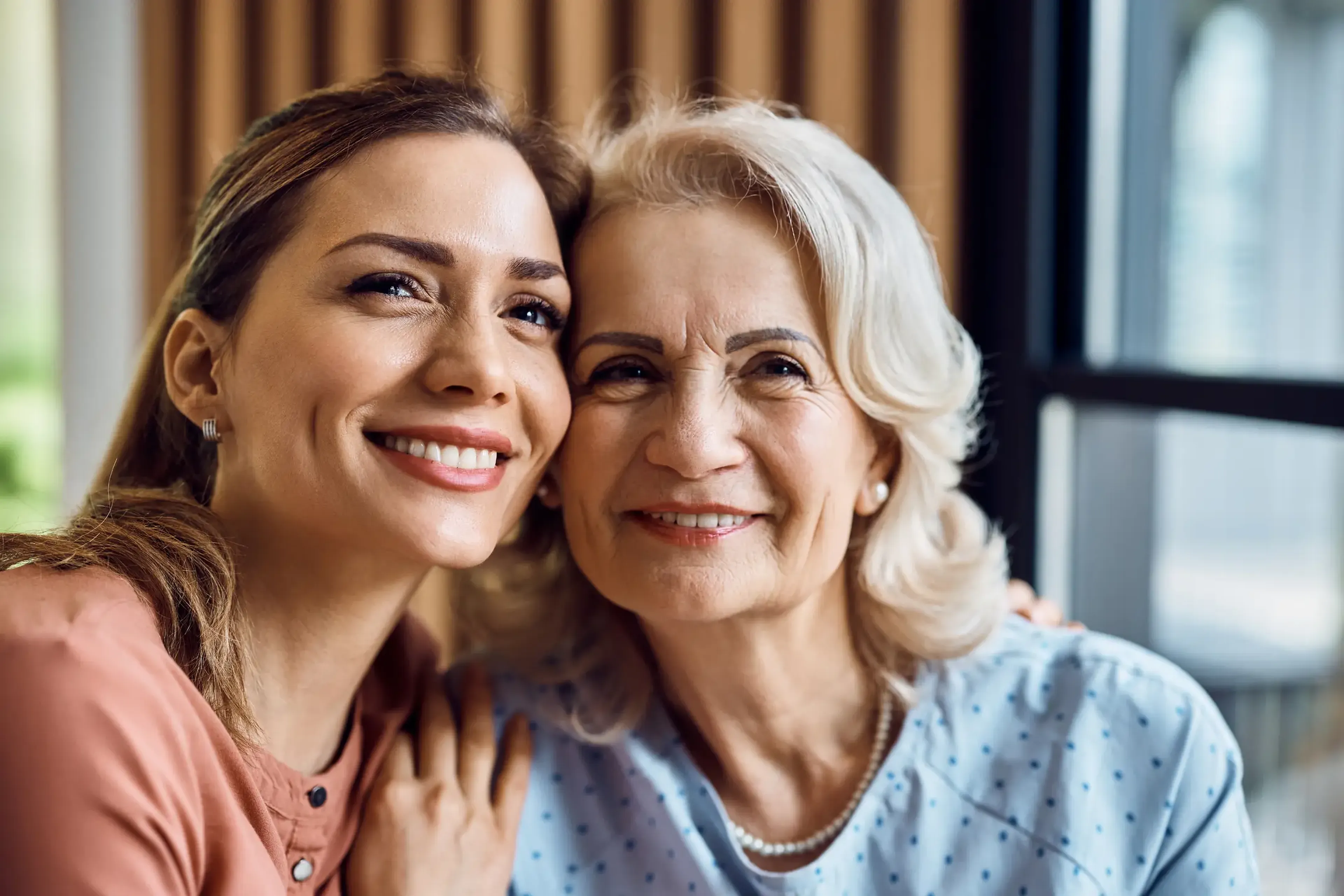 Two women smiling warmly, one younger with brown hair, the other older with gray hair, indoors near a window.