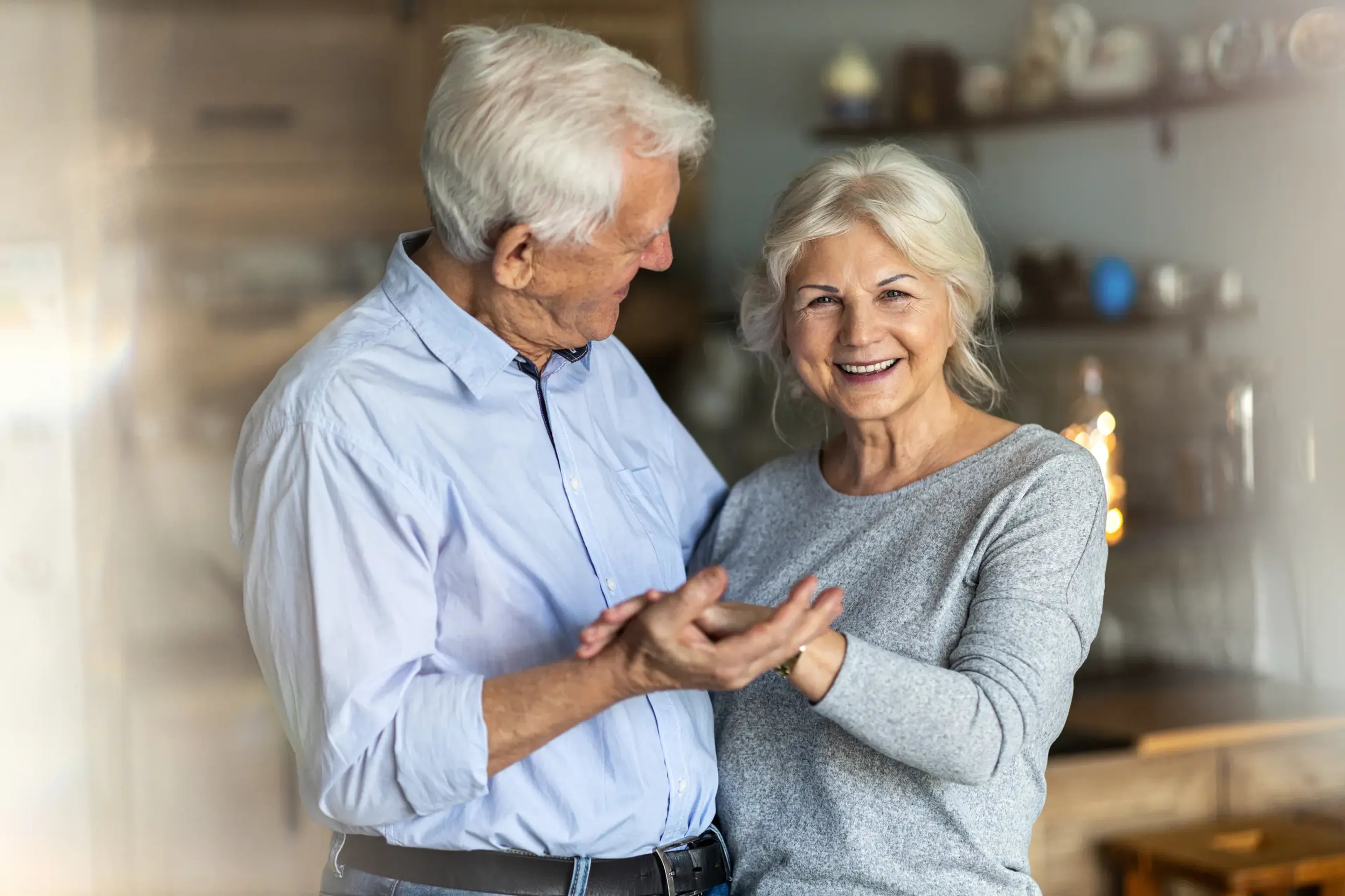 Elderly couple smiling and holding hands in a cozy kitchen, with the woman looking at the camera and the man gazing at her affectionately.