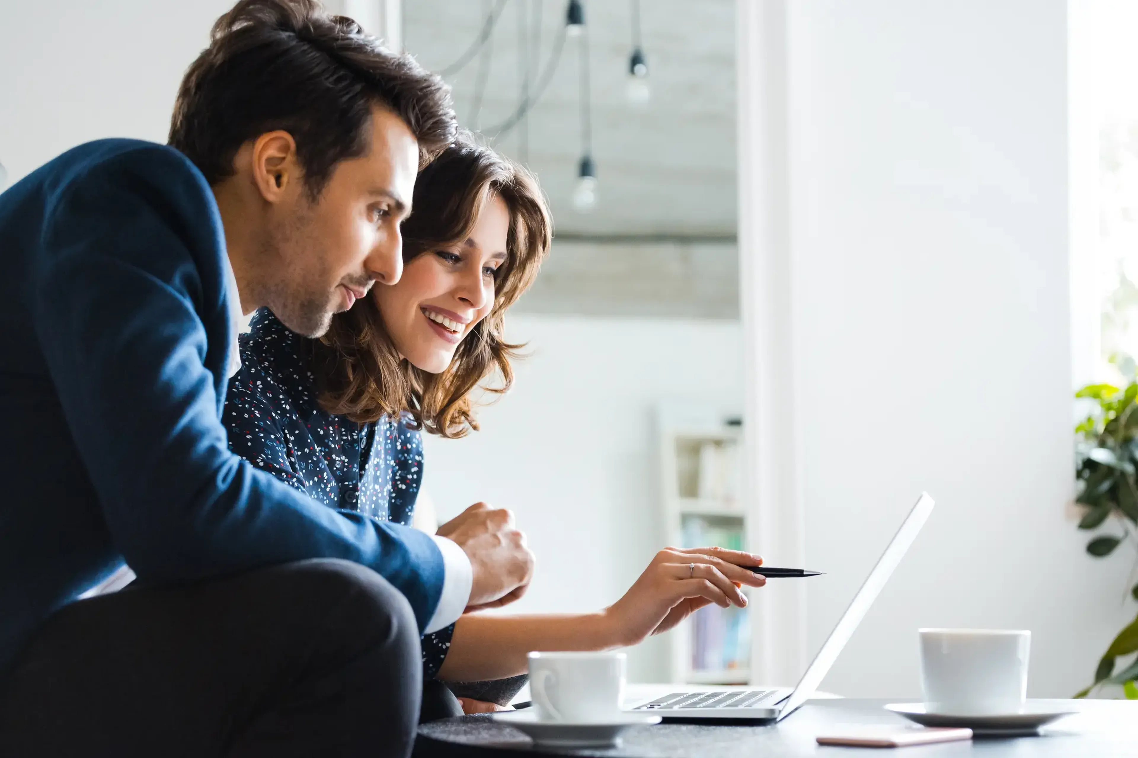 A man and woman smiling while looking at a laptop screen, sitting at a table with coffee cups in a bright room.