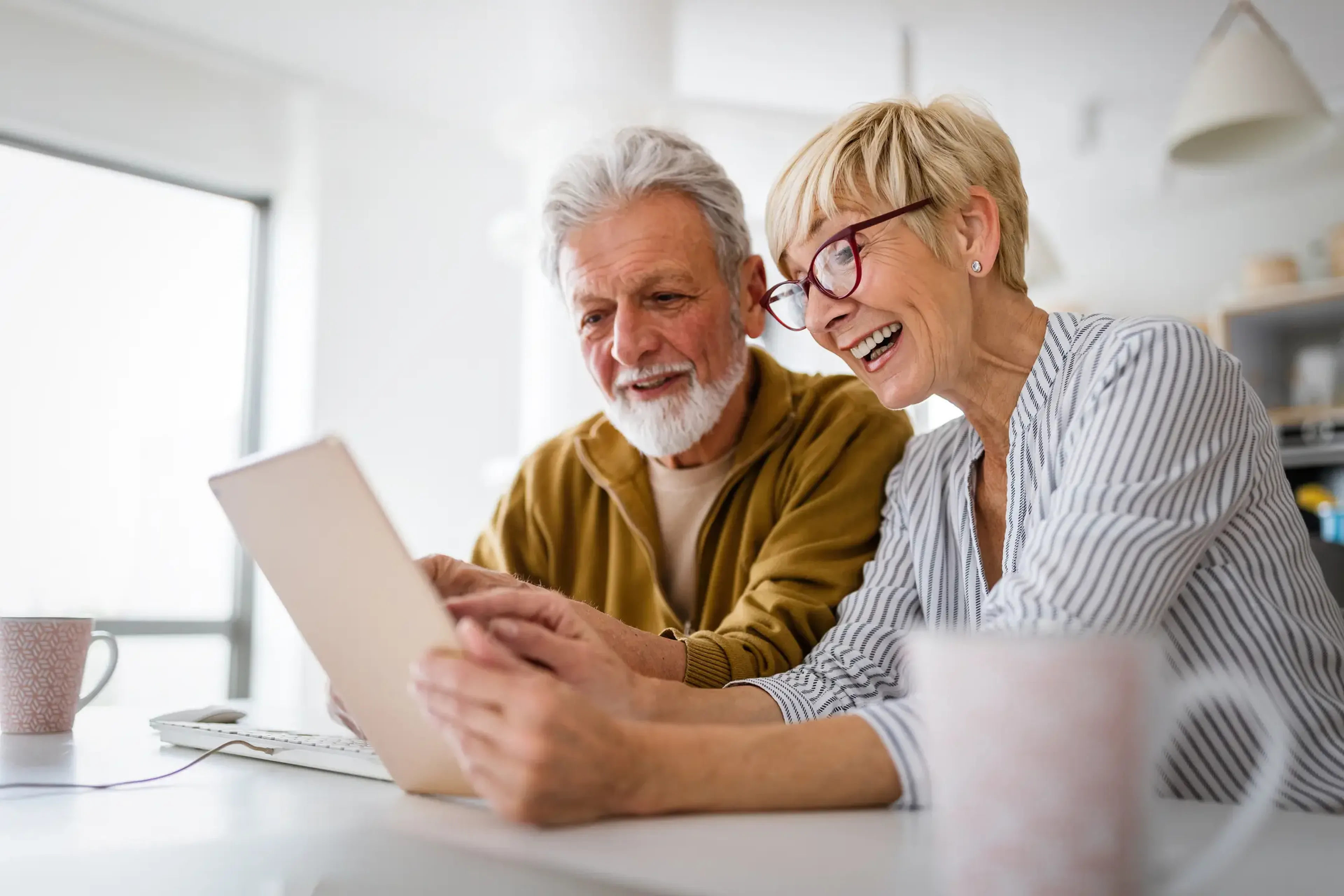Elderly couple smiling and looking at a tablet together in a bright room, with two coffee mugs on the table.