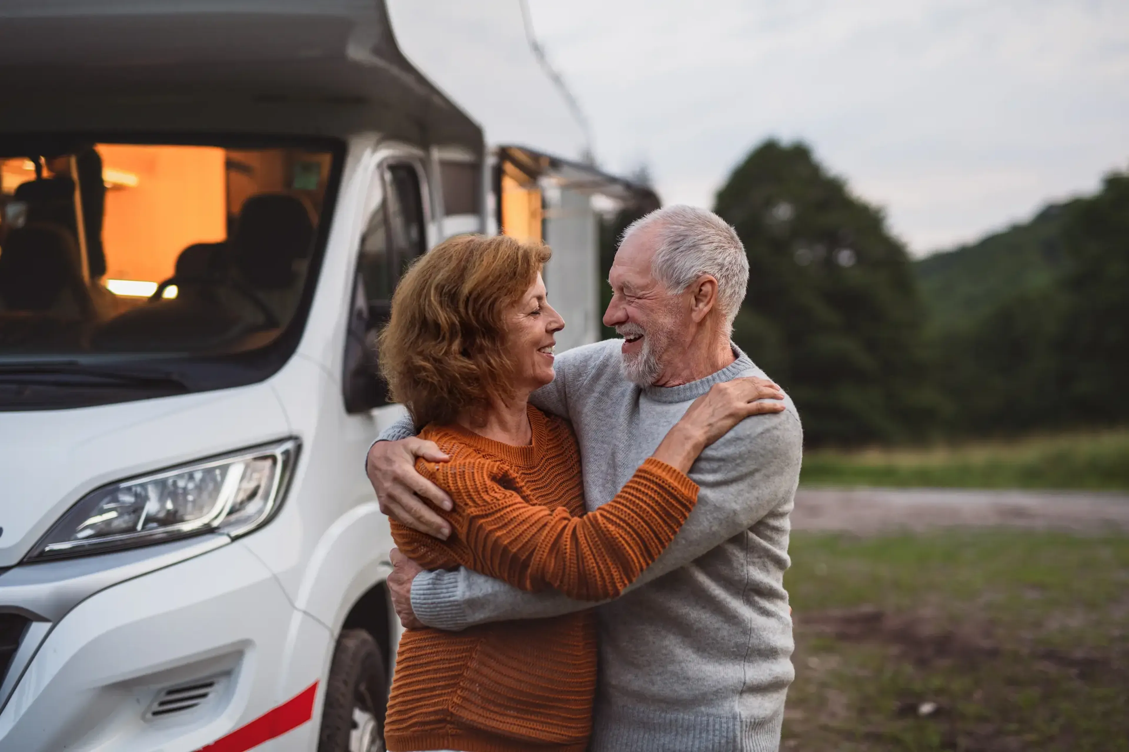 Elderly couple embracing and smiling in front of a camper van, parked in a scenic outdoor setting with trees in the background.