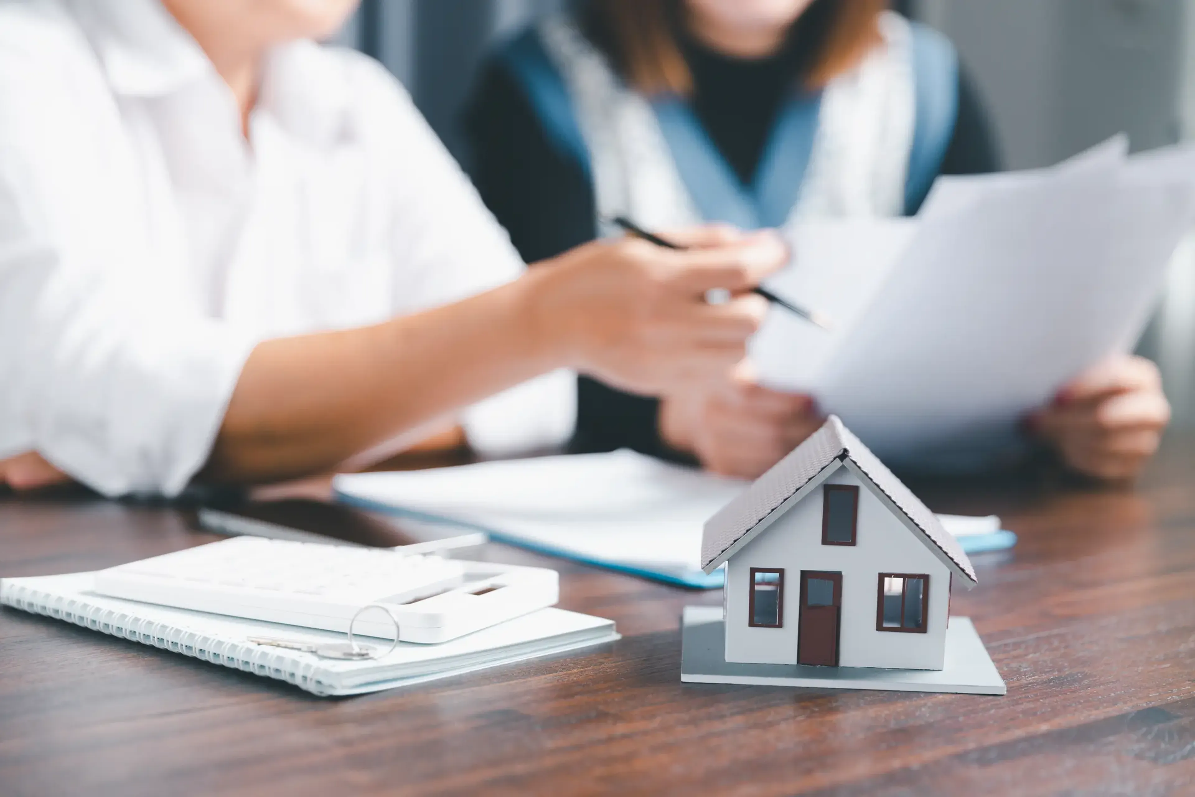Two people discussing documents at a table with a small house model, notebook, and calculator in focus.