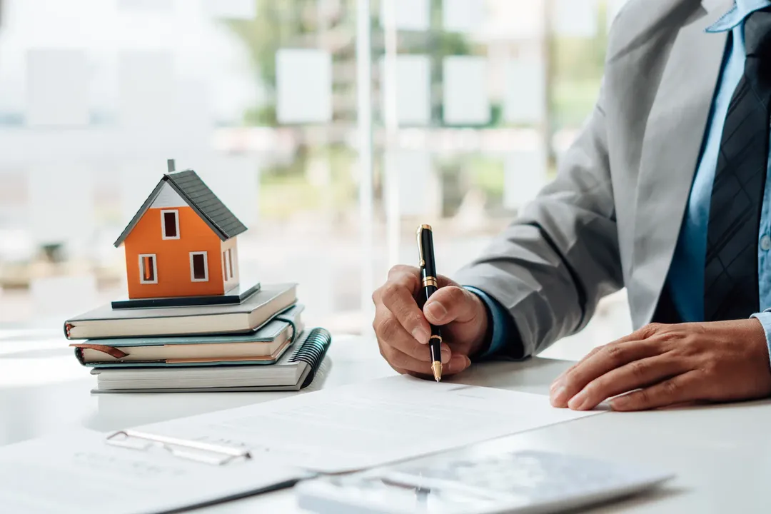 Person signing documents at a desk with a small house model on stacked books, symbolizing real estate or mortgage.