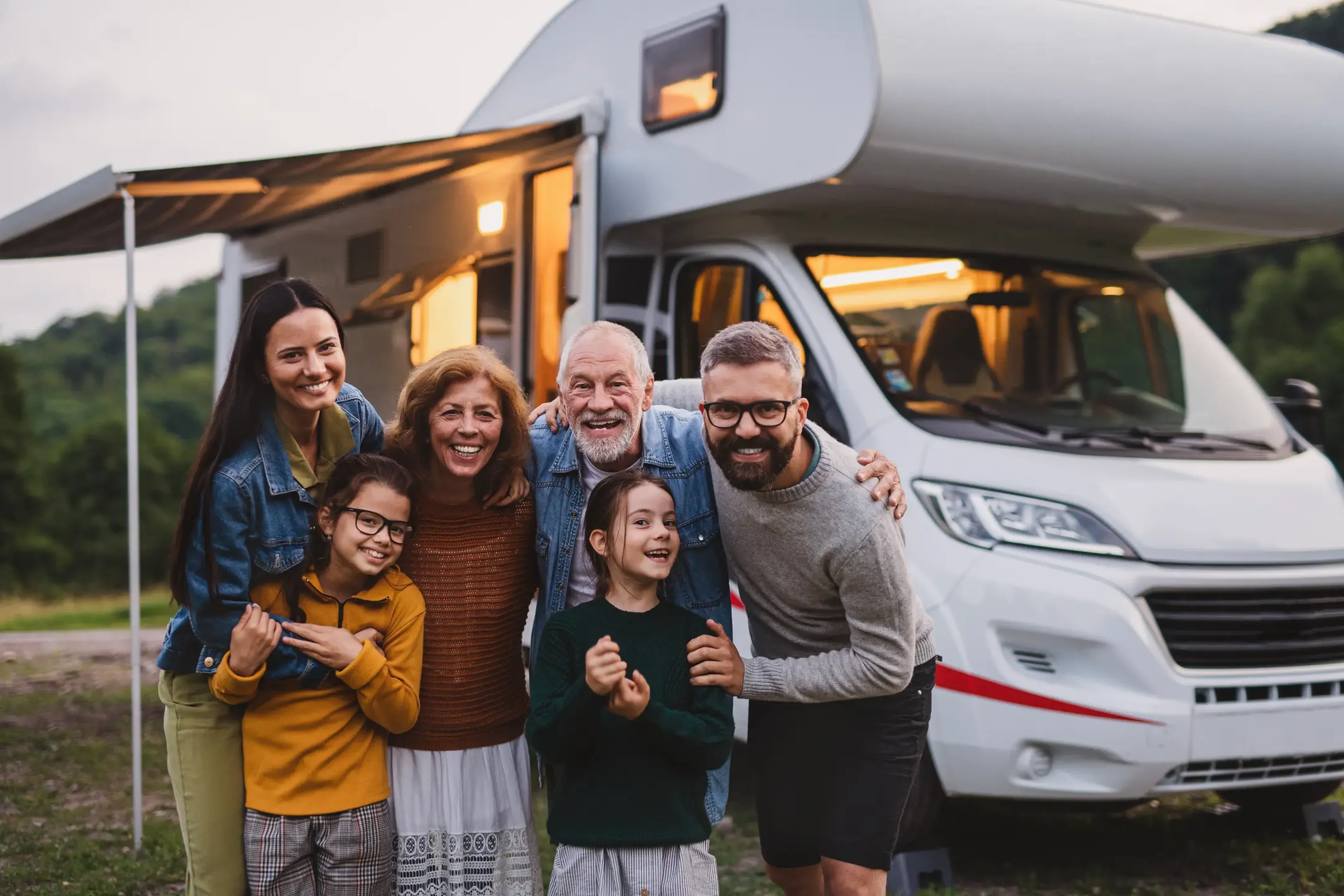 Family smiling and posing in front of a camper van, parked outdoors. Two adults, two children, and two seniors are enjoying their time together.