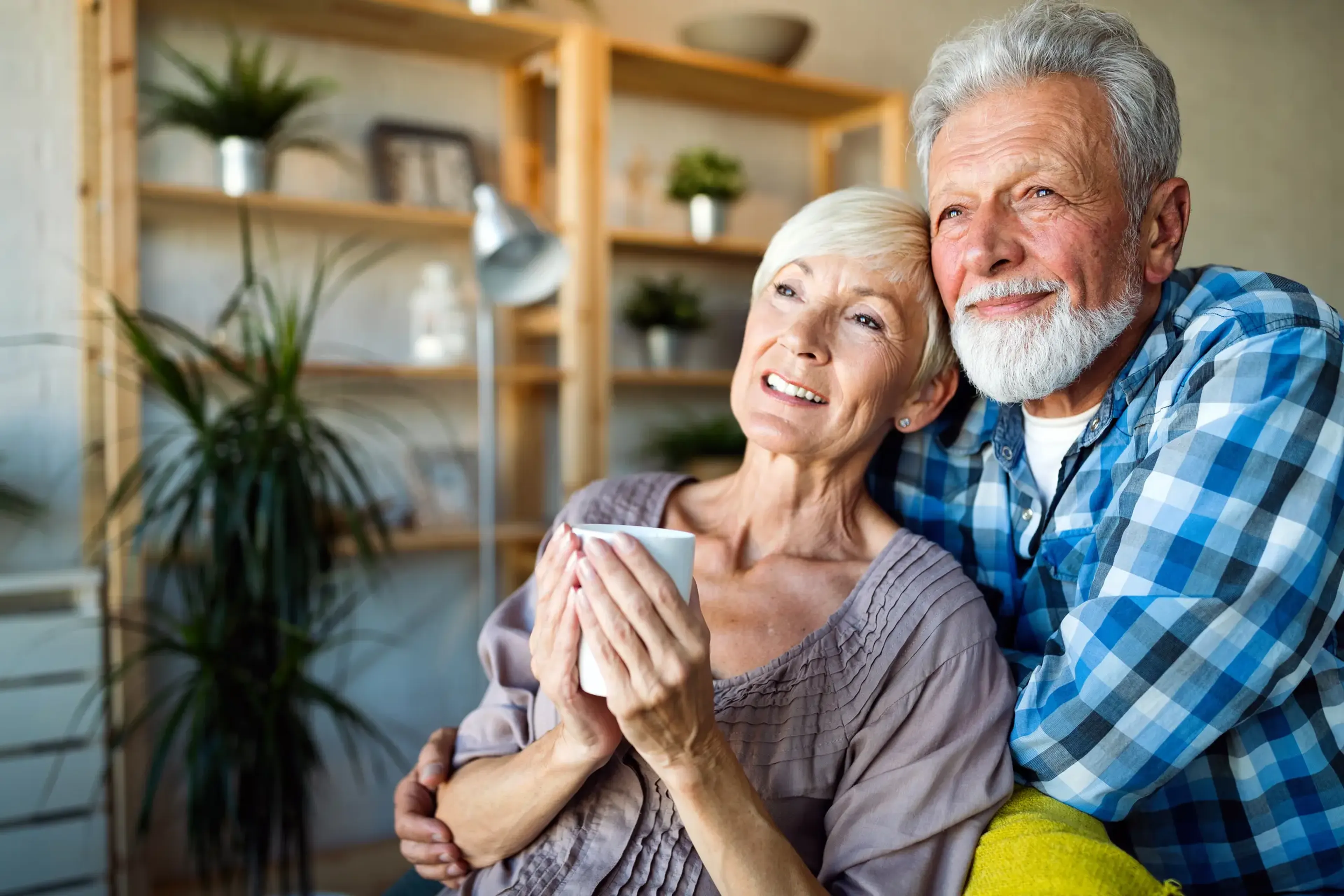 Elderly couple smiling while sitting together on a couch, with the woman holding a mug. Shelves with plants are in the background.