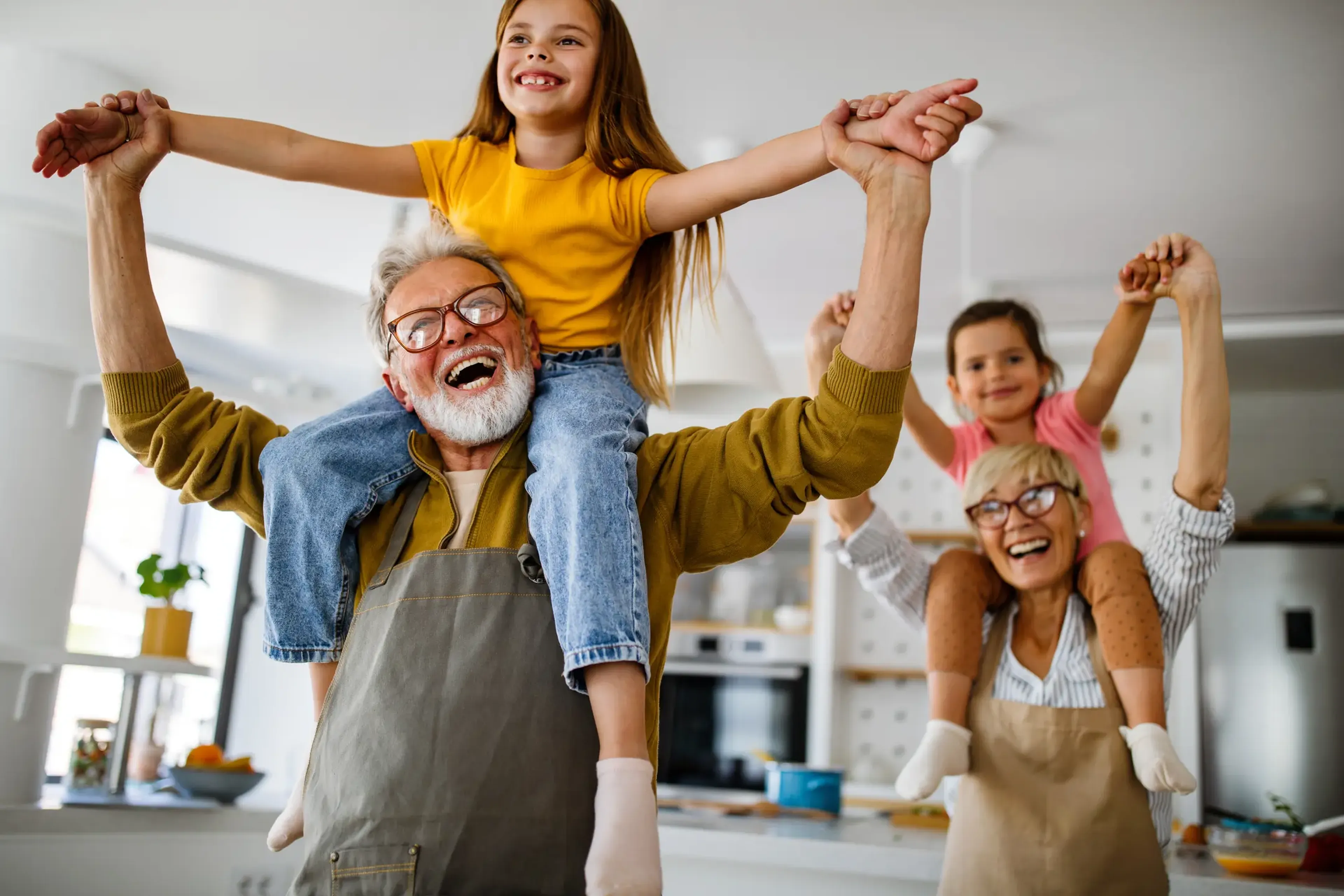 Two joyful children sit on the shoulders of smiling grandparents in a bright kitchen, holding hands and laughing together.