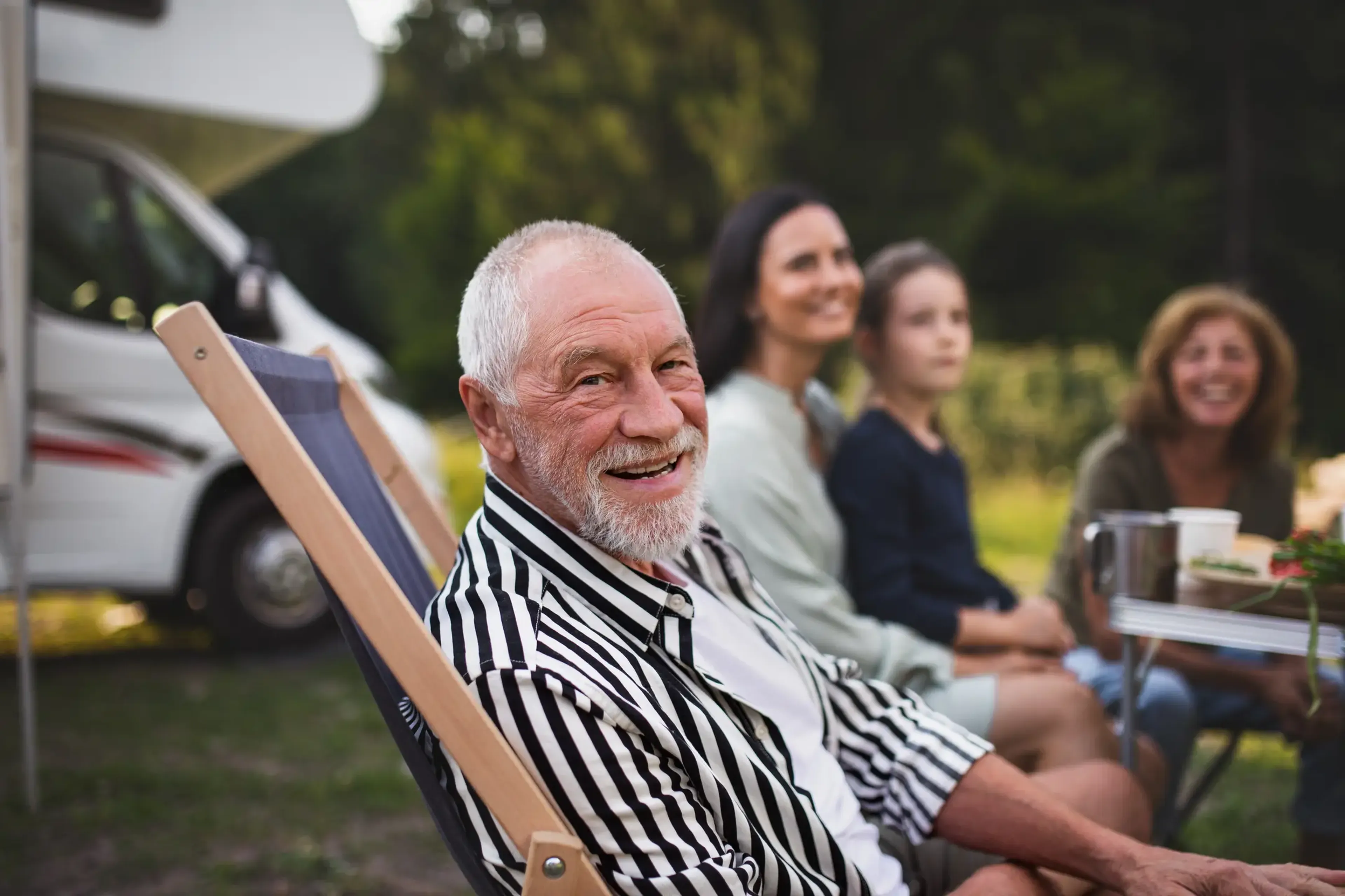 Elderly man smiling in a striped shirt, sitting outdoors with three people near a camper, surrounded by greenery.