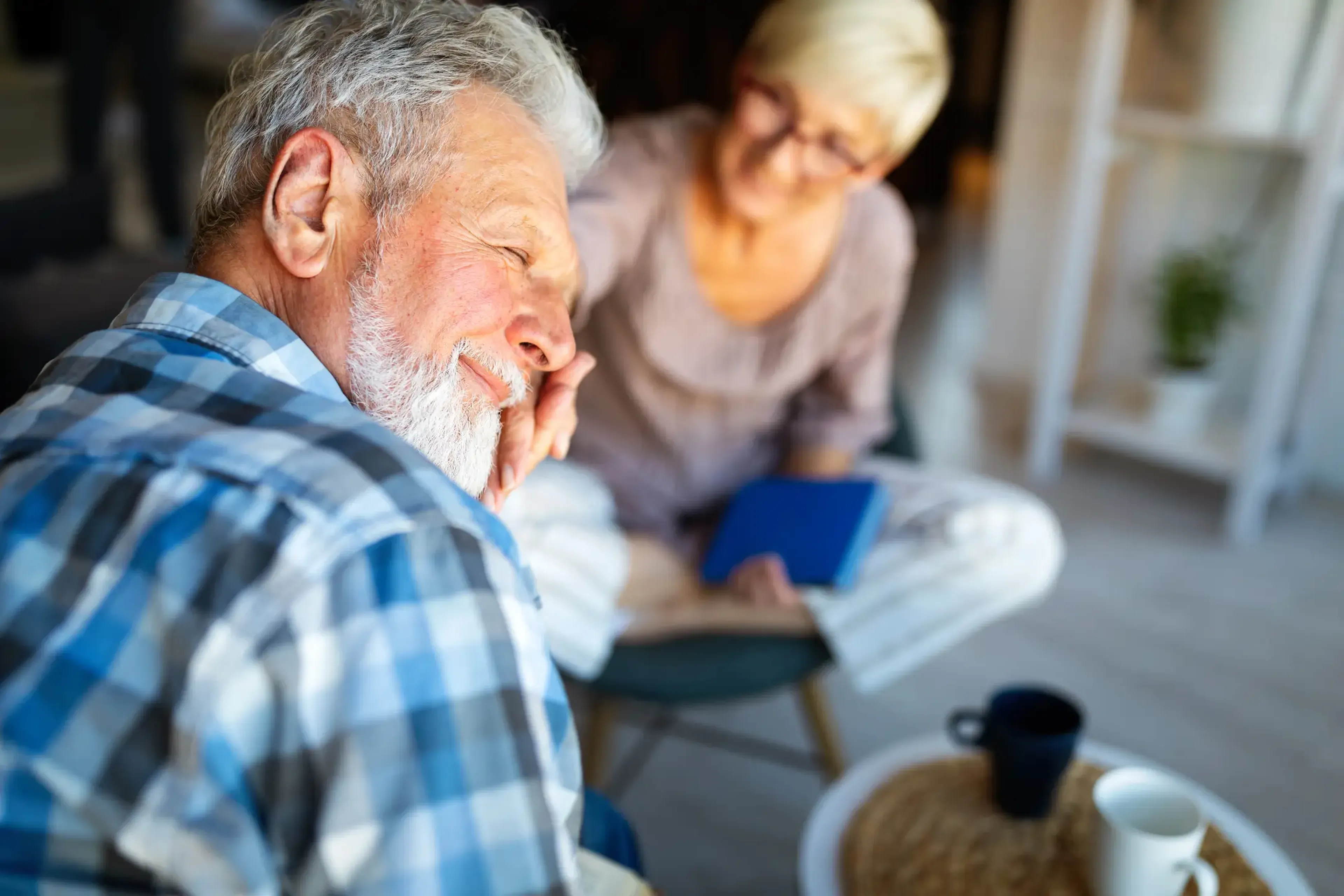 Elderly couple smiling warmly at each other, the woman holding a book, sitting in a cozy room with a table and cups.