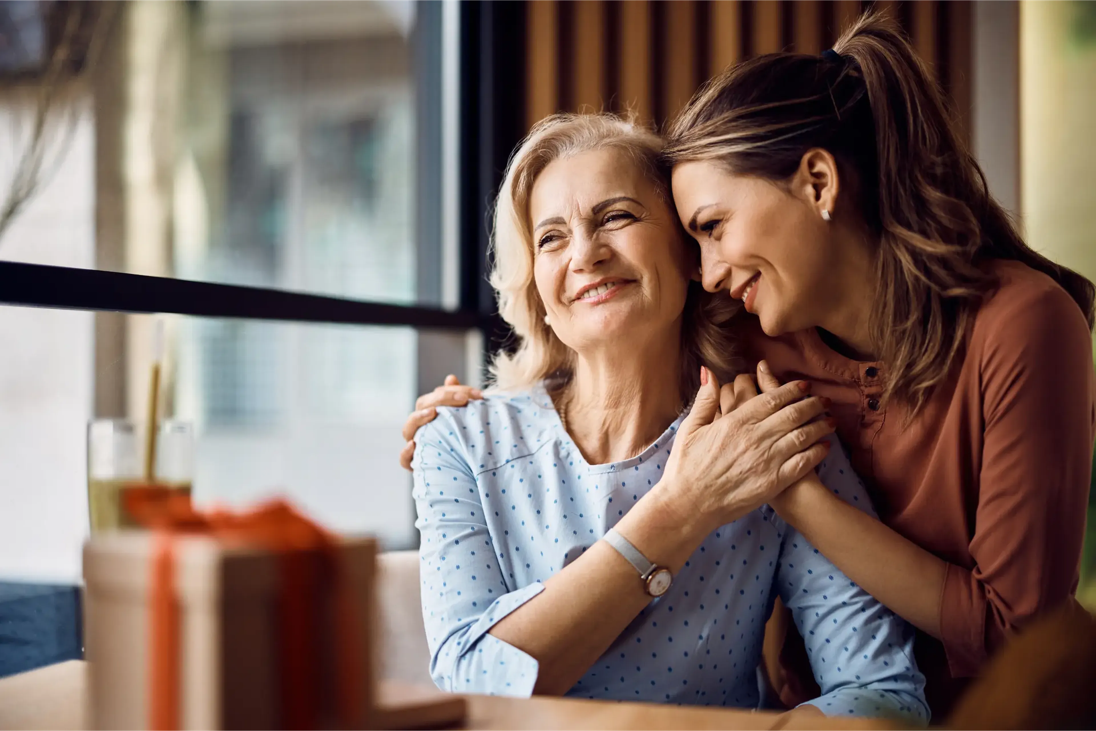 A smiling older woman and a younger woman embrace affectionately at a table with a gift box, near a large window.