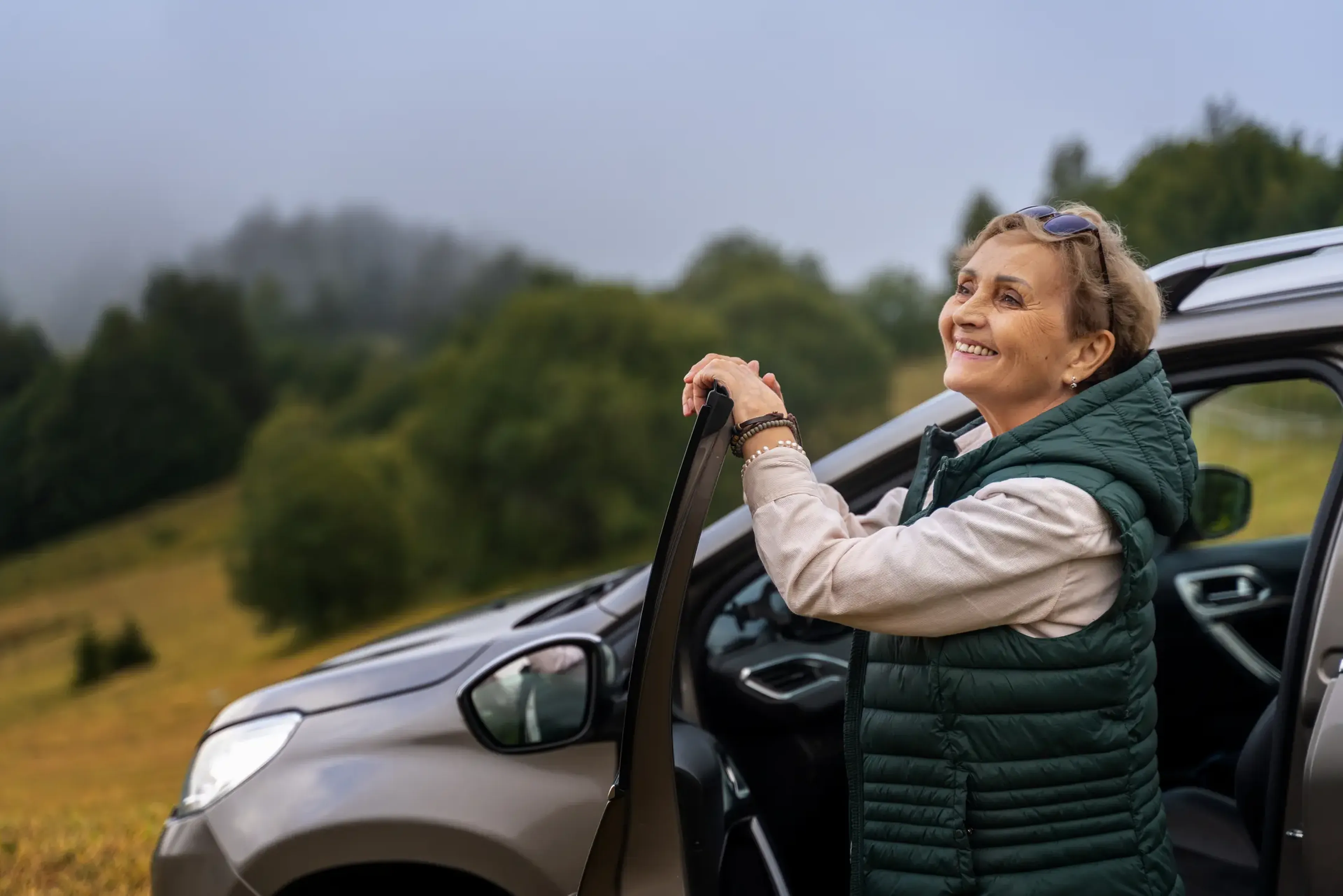 Smiling woman in a green vest stands by an open car door, enjoying a misty, lush green landscape.