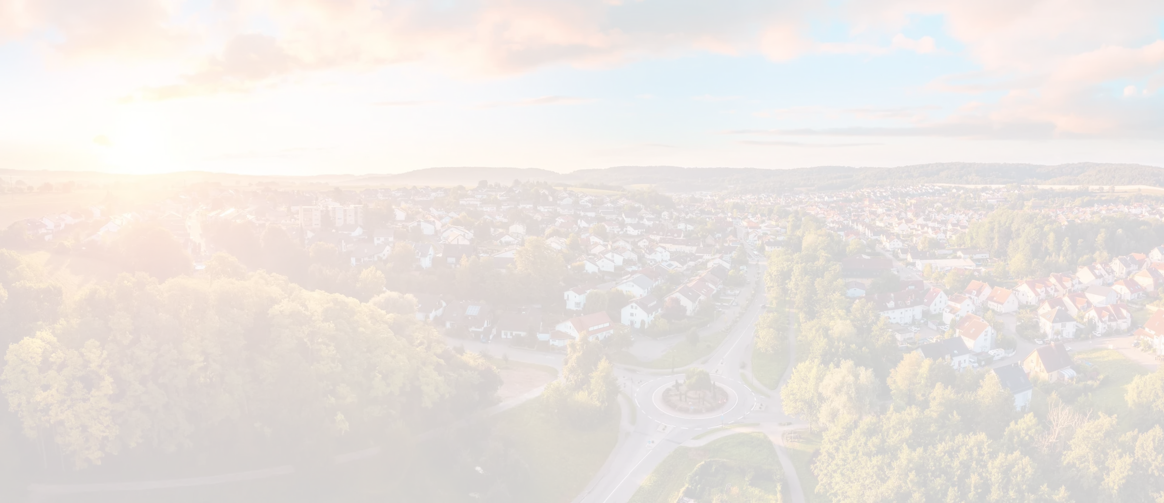 Aerial view of a suburban neighborhood at sunset, with houses, roads, lush greenery, and a vibrant sky with clouds.