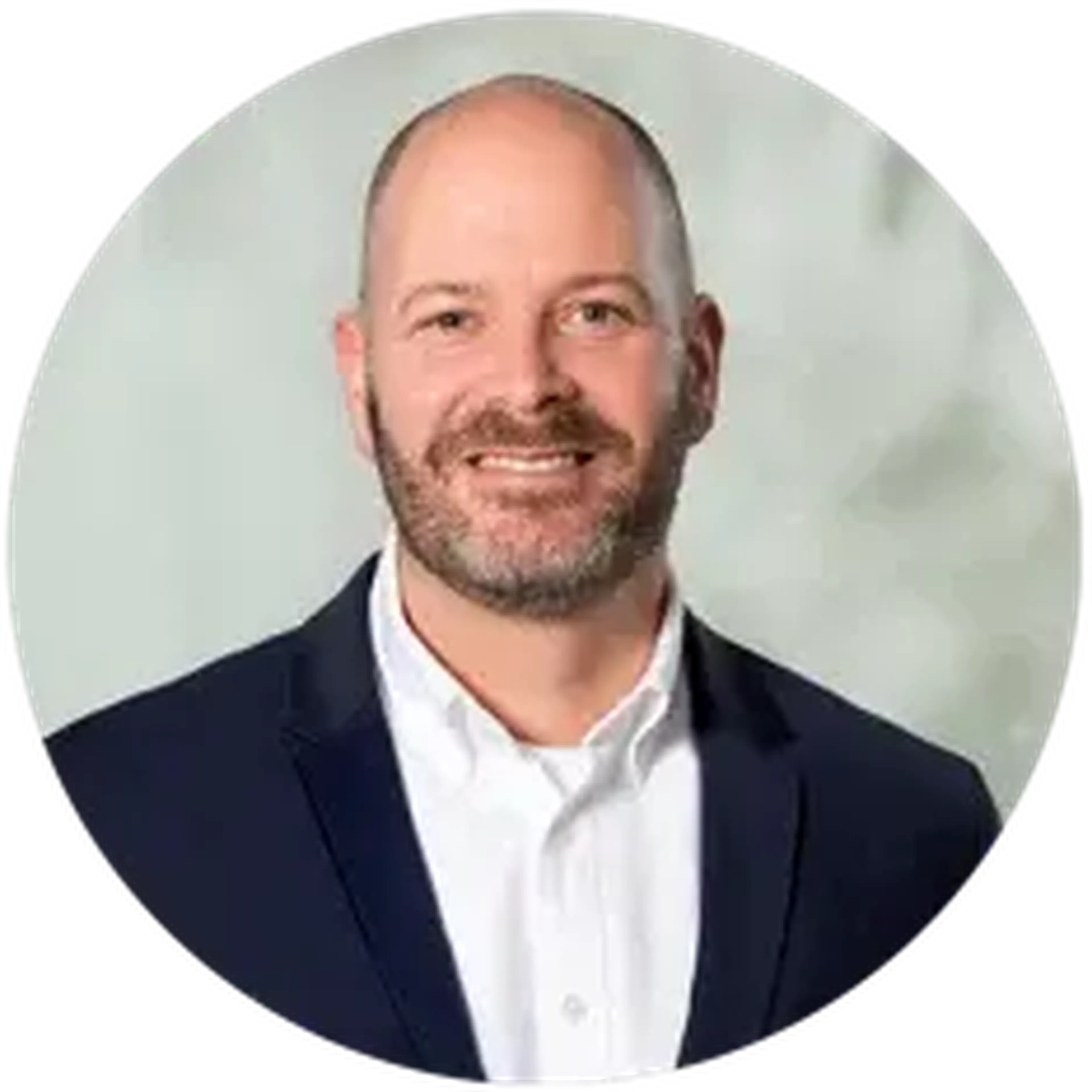 A man with a beard and a shaved head, wearing a dark suit and white shirt, smiles against a light gray background.