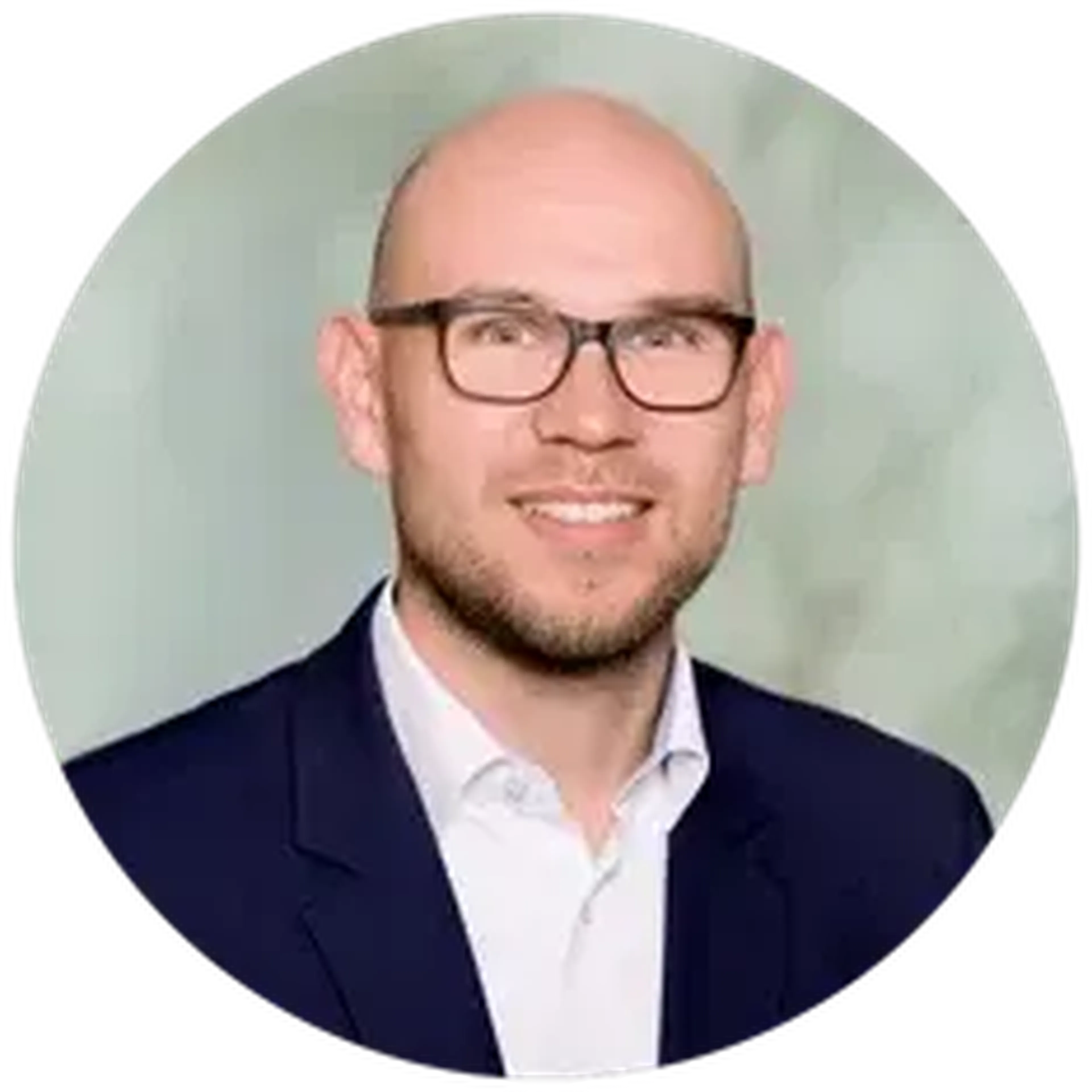 A smiling man with glasses, a bald head, and a beard, wearing a dark suit and white shirt, against a light, blurred background.