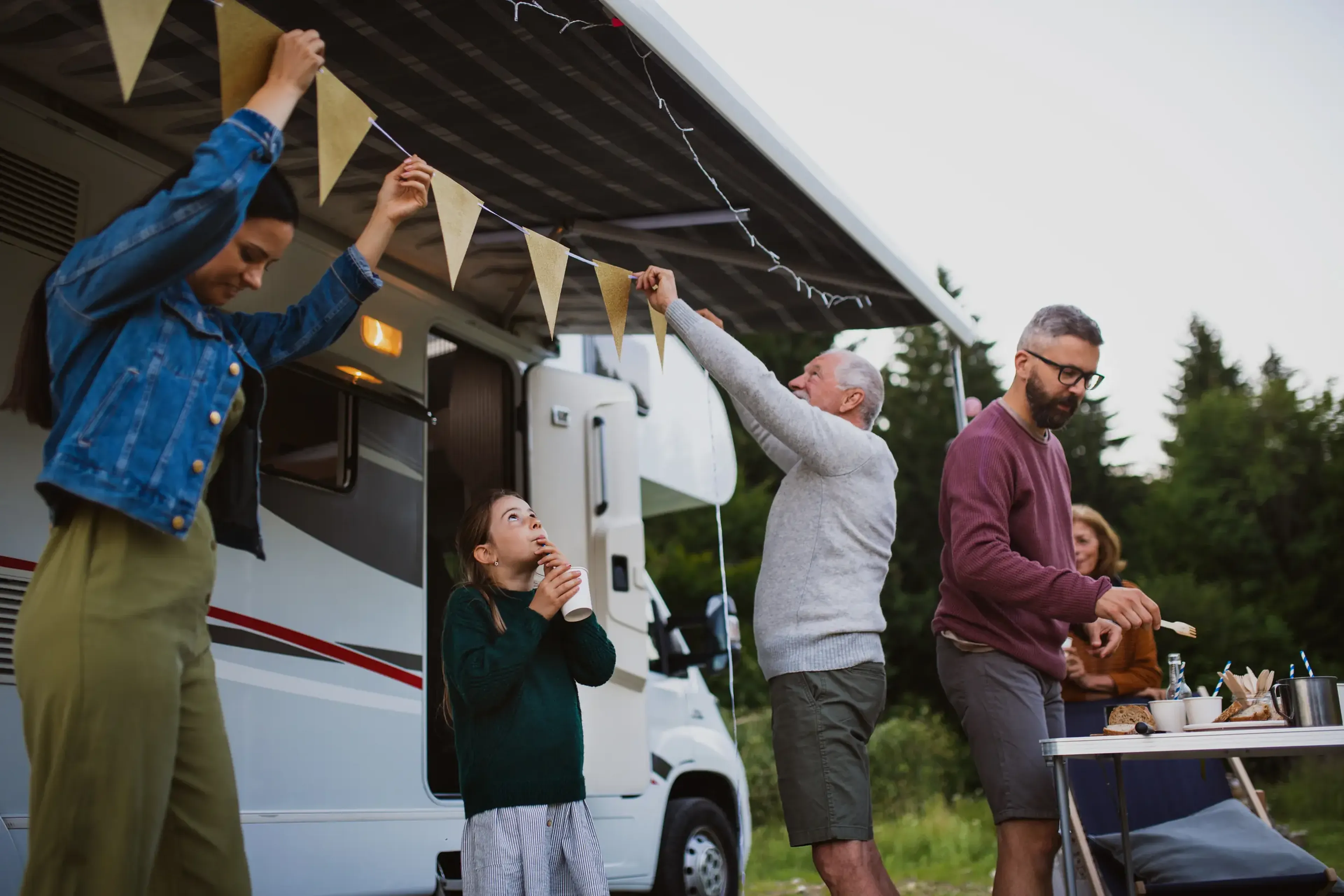Family decorating a camper van with bunting. A man and child hang flags, while others prepare food on a table. Trees in the background.