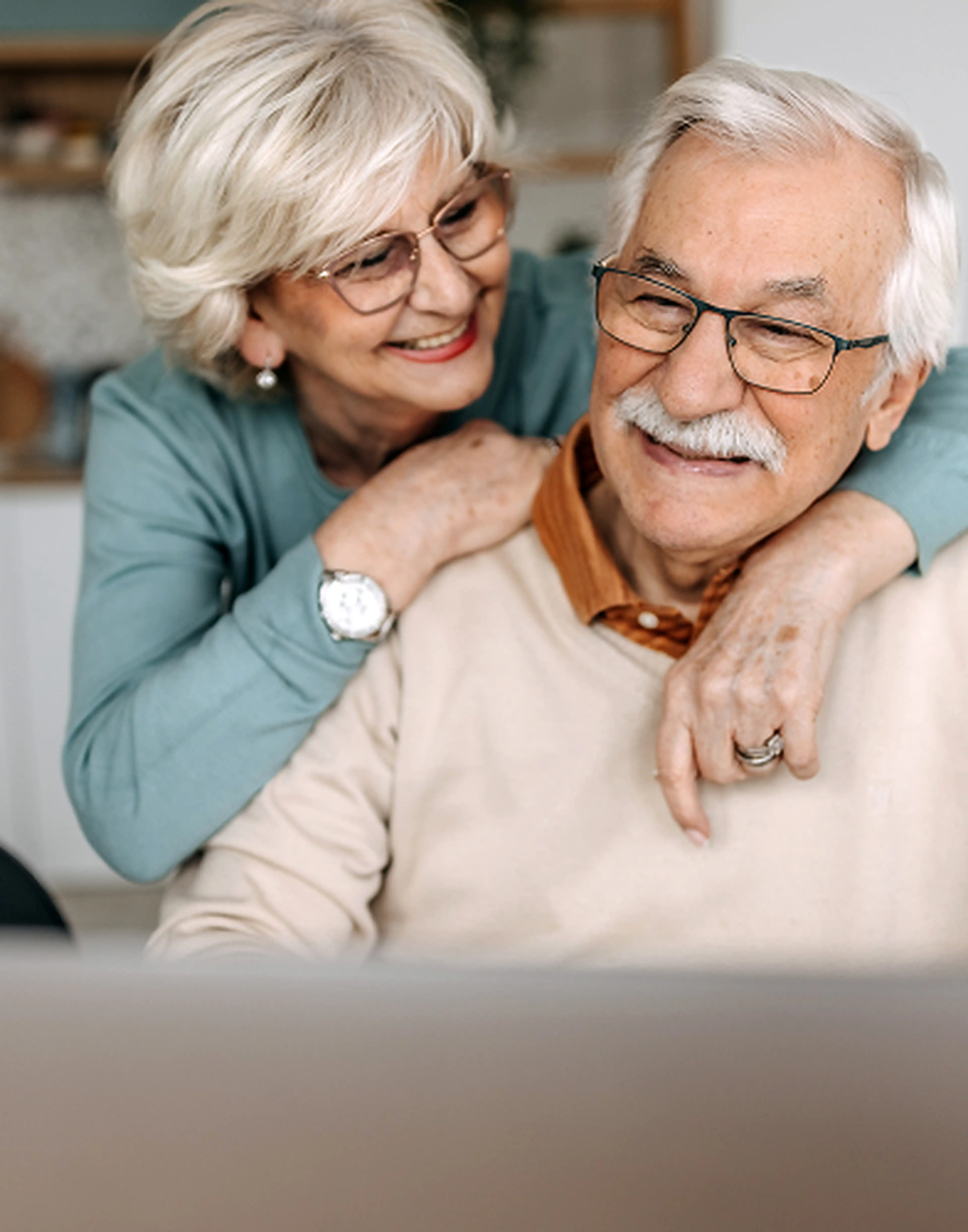 Senior woman smiling as a younger woman embraces her, sharing a warm, affectionate moment indoors.