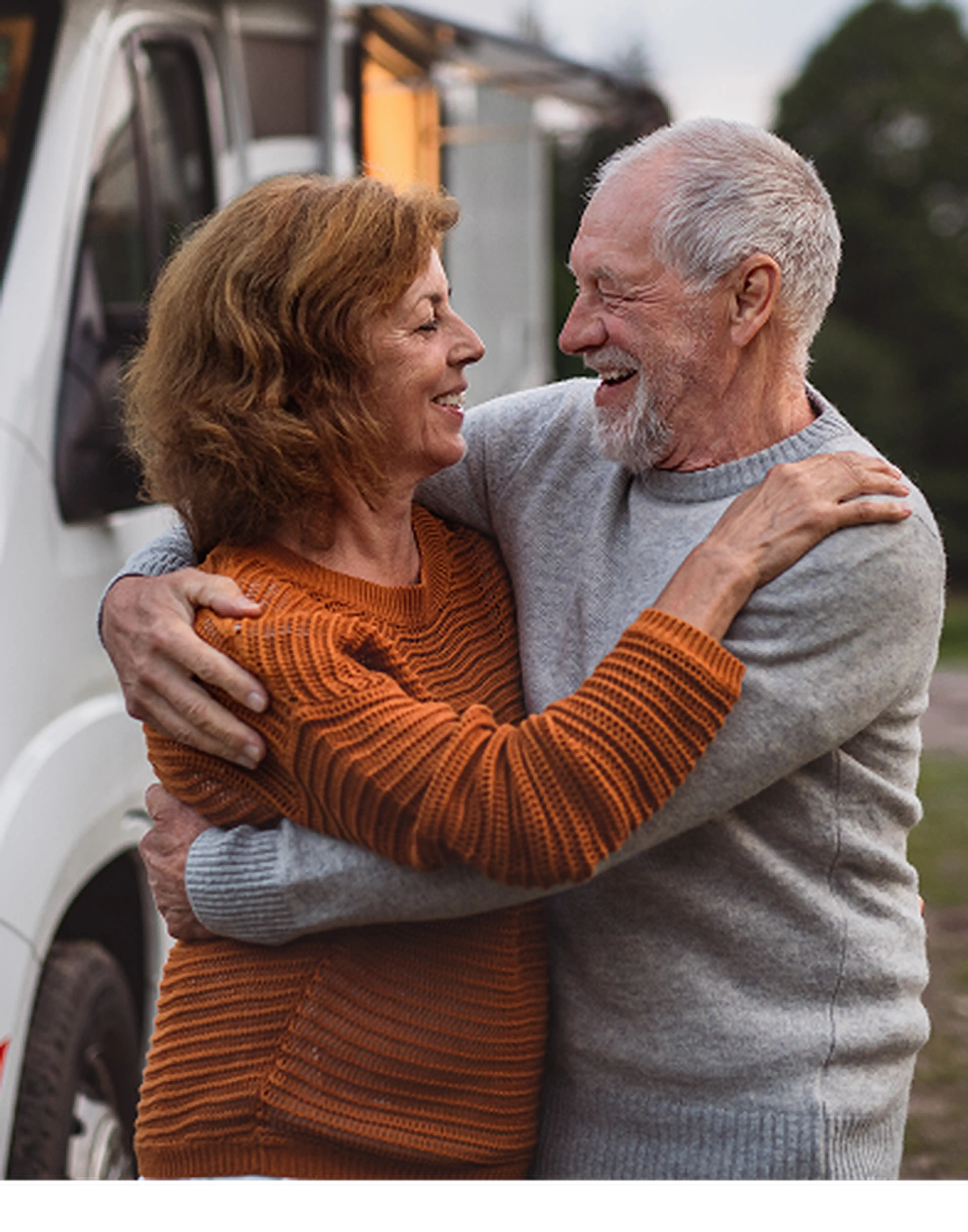 Smiling older couple embracing each other outdoors in front of a white camper van, sharing a joyful moment