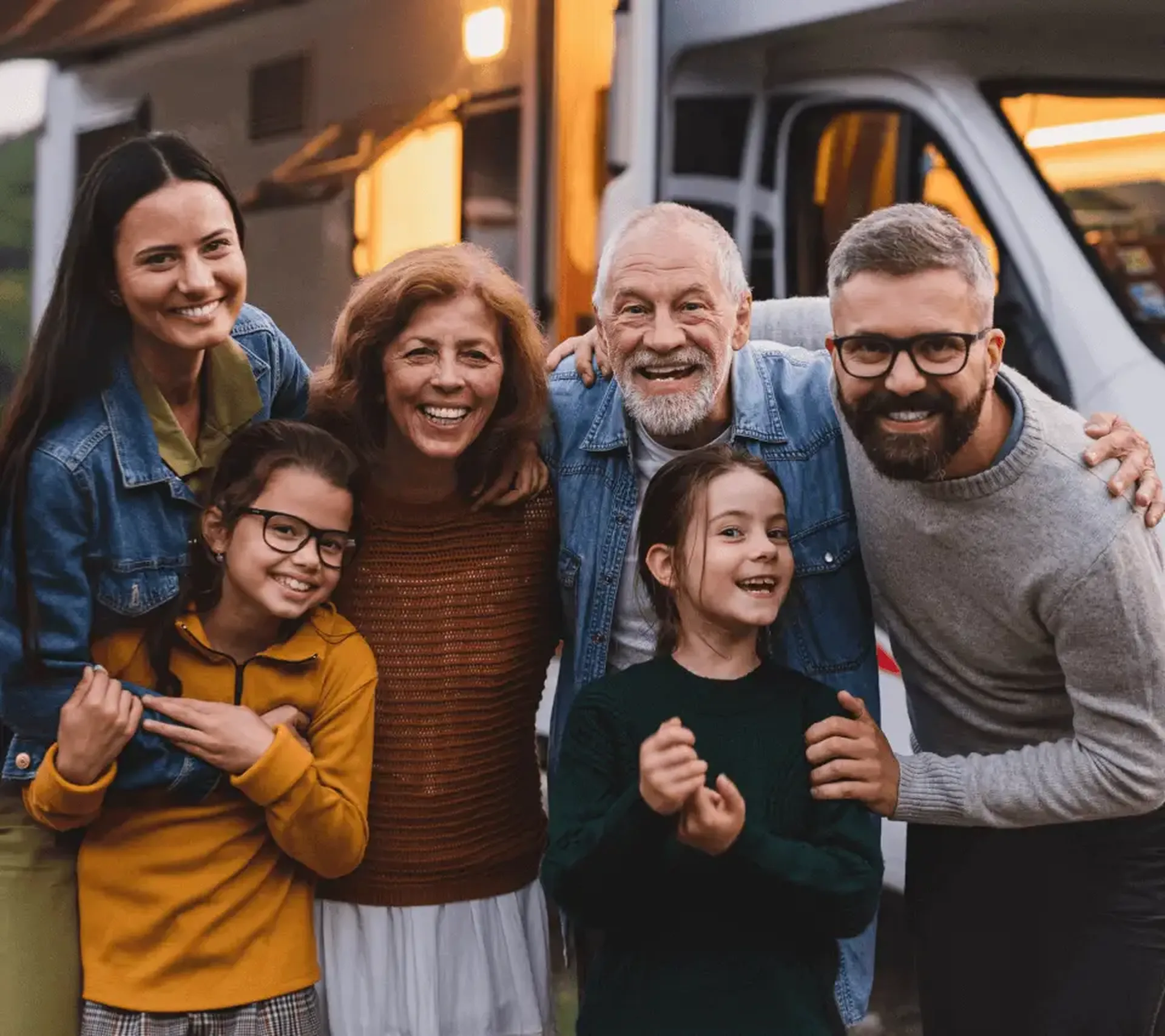 A joyful family of six, spanning three generations, stands together smiling in front of a camper van, enjoying an outdoor gathering.
