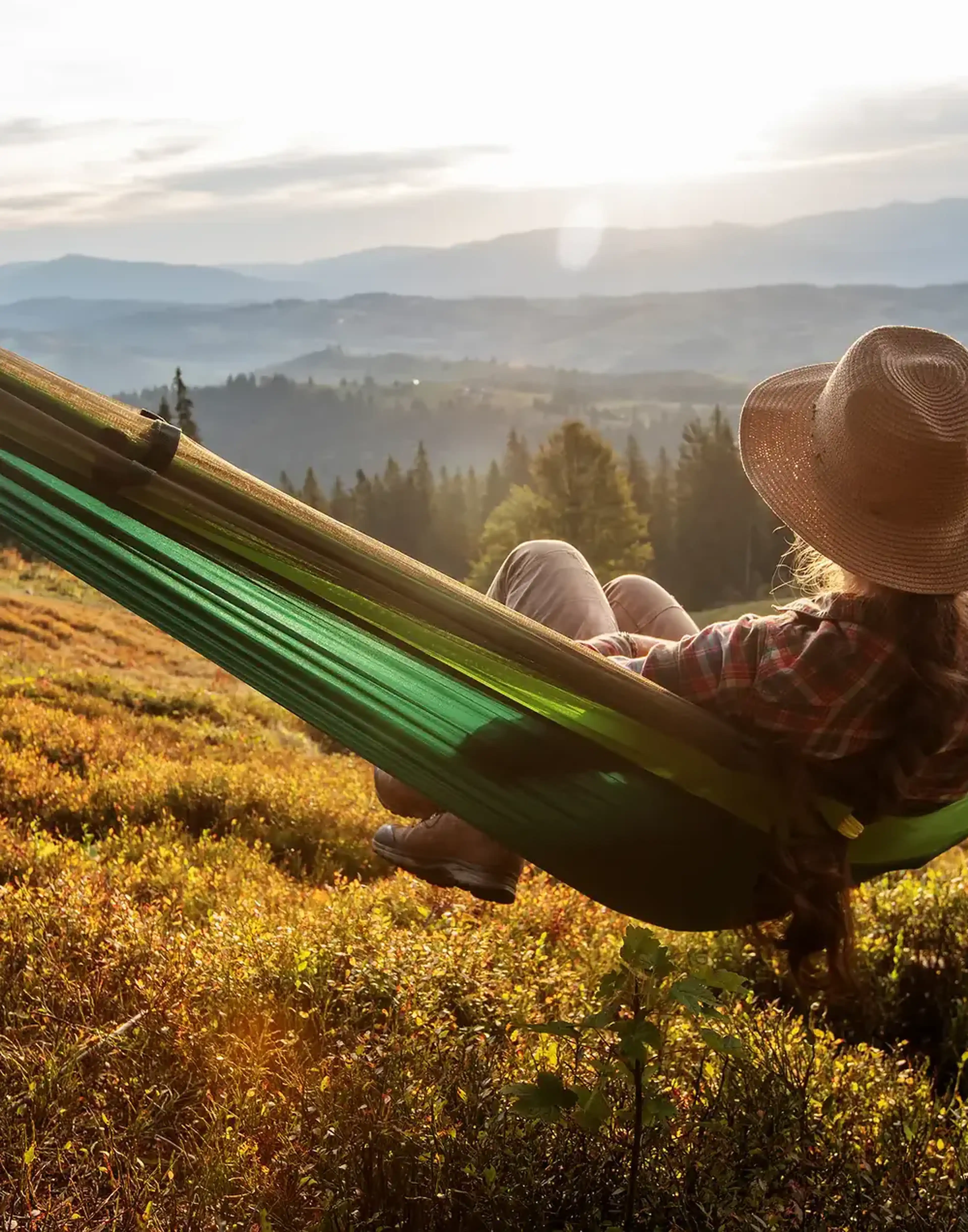 Person in a hat relaxing in a green hammock on a hillside, overlooking a scenic view of mountains and forests at sunset.