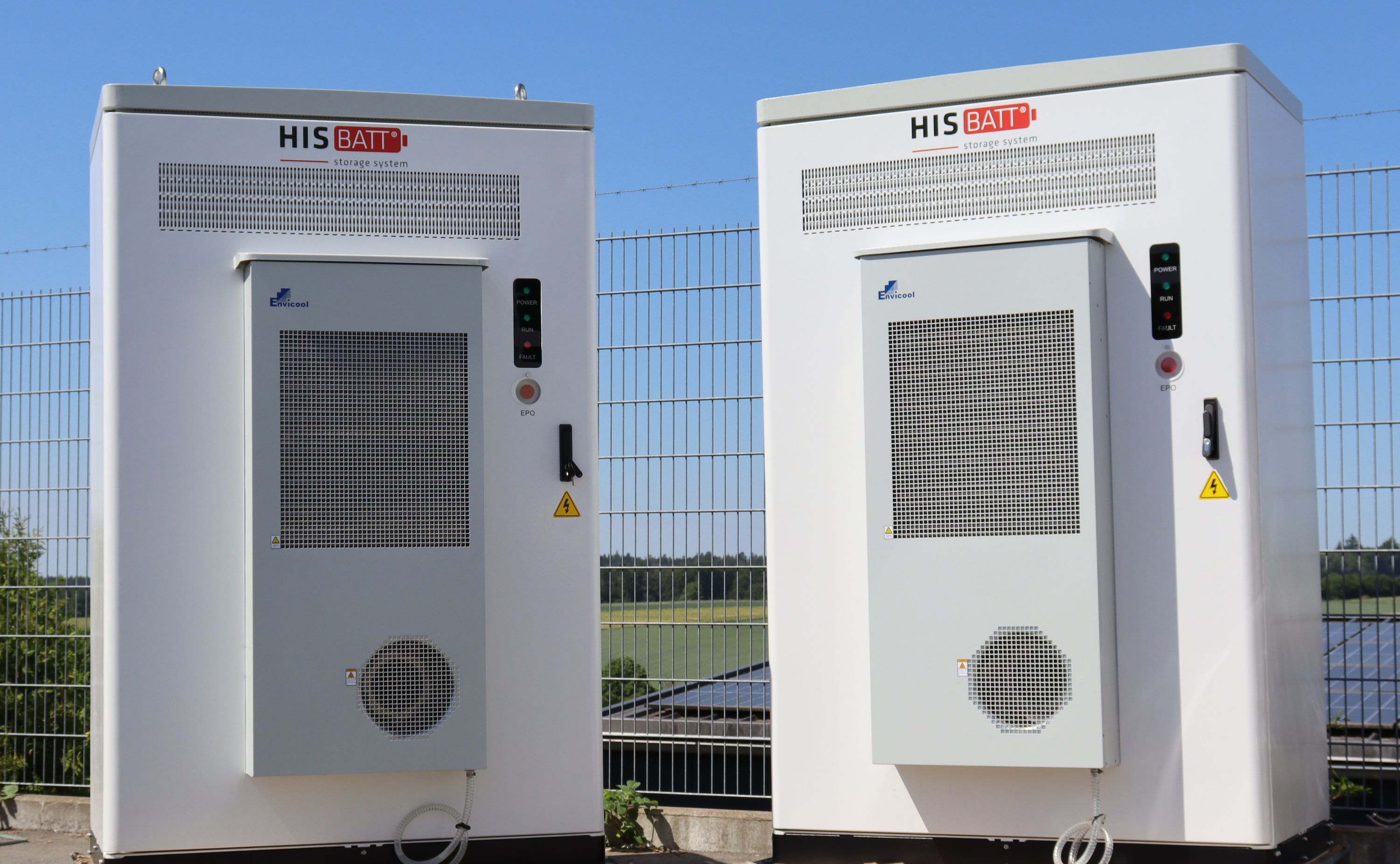 Two white HIS BATT storage systems with vented panels and control units, set against a blue sky and metal fence.