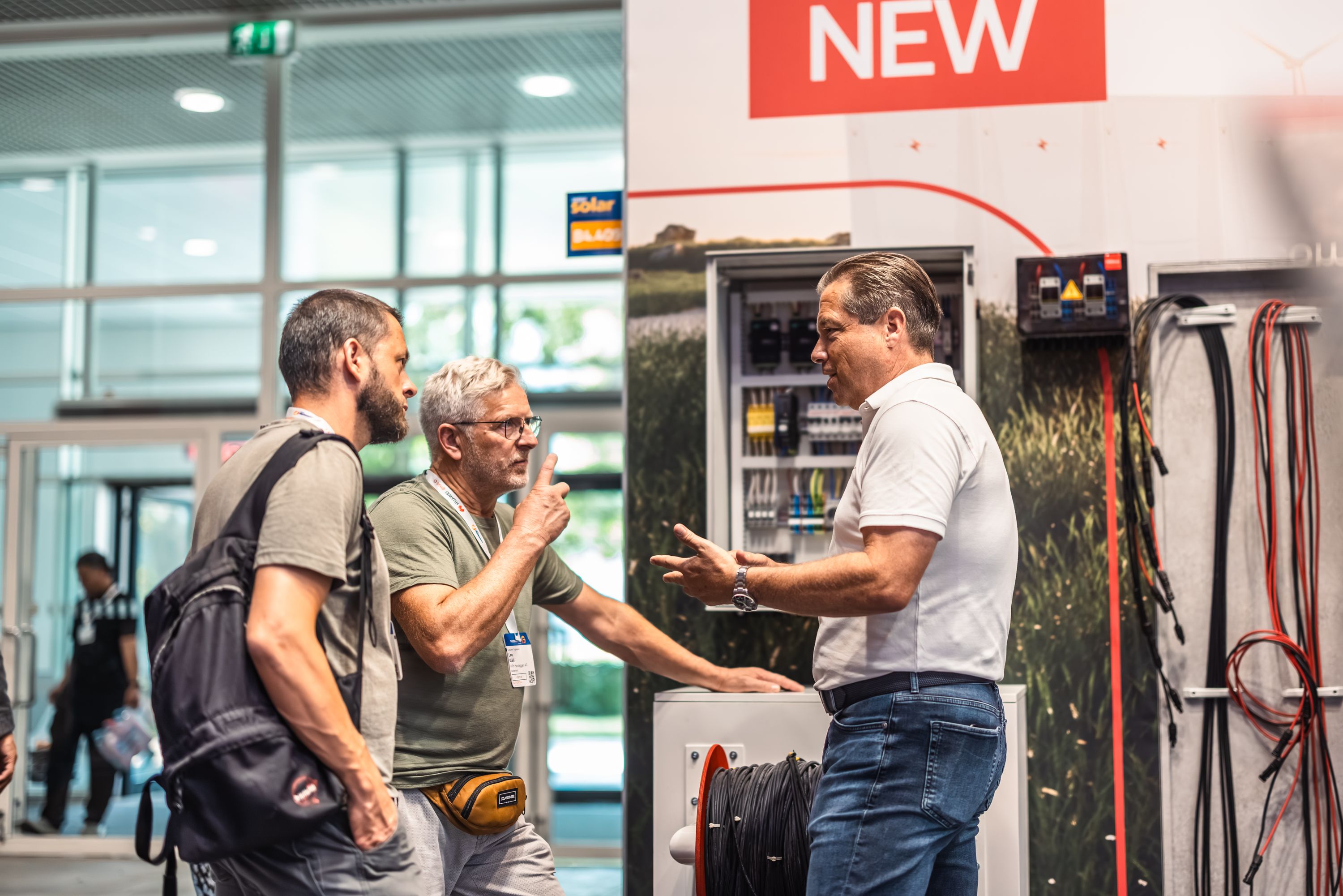 Three men engaged in a discussion near an open electrical panel at a trade show or exhibition, with cables and a sign reading "NEW" in the background.