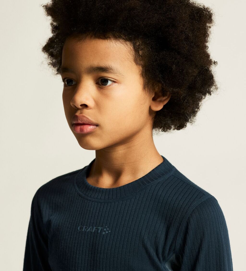 Lite barn med krøllete hår iført en mørk ribbet skjorte. Barnet ser til siden mot en ensfarget bakgrunn.