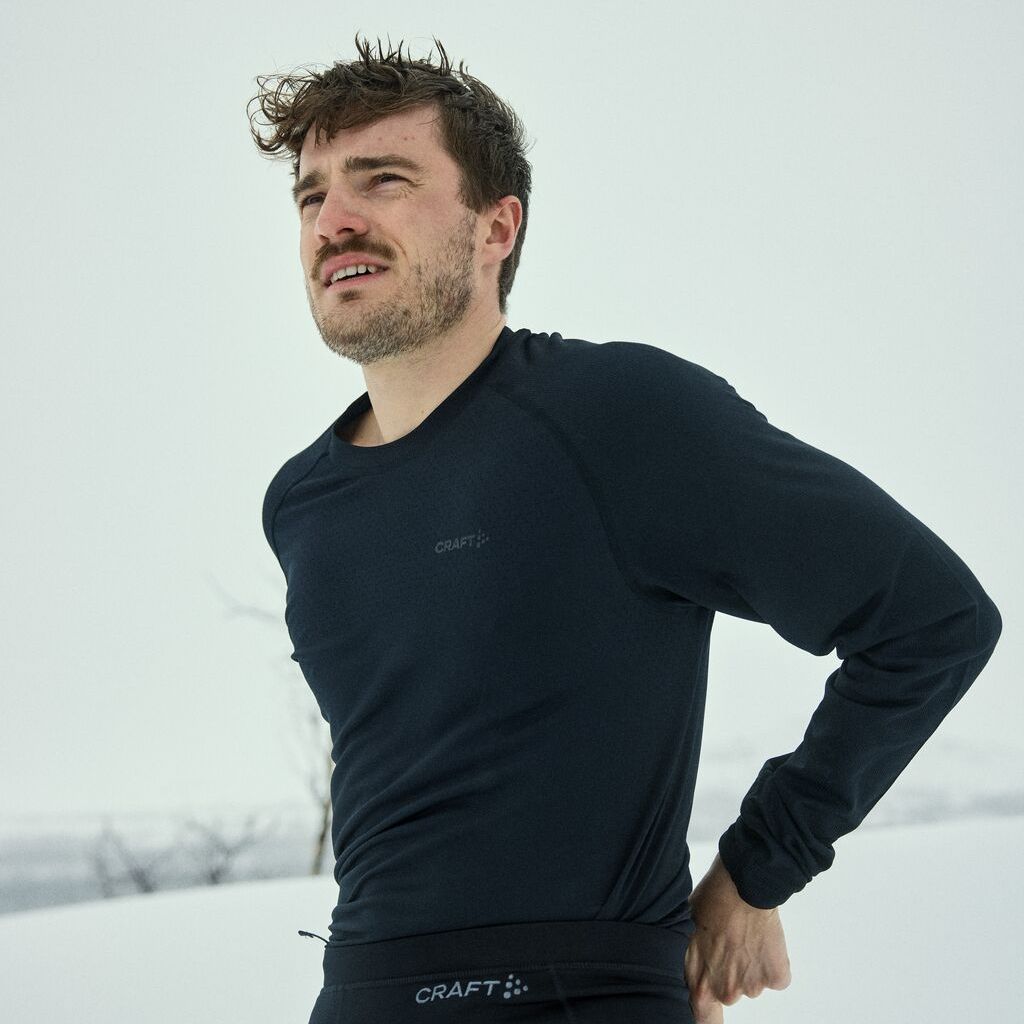 En mann i en svart langermet genser står utendørs i et snødekt landskap og retter på klærne sine, med et ettertenksomt uttrykk.