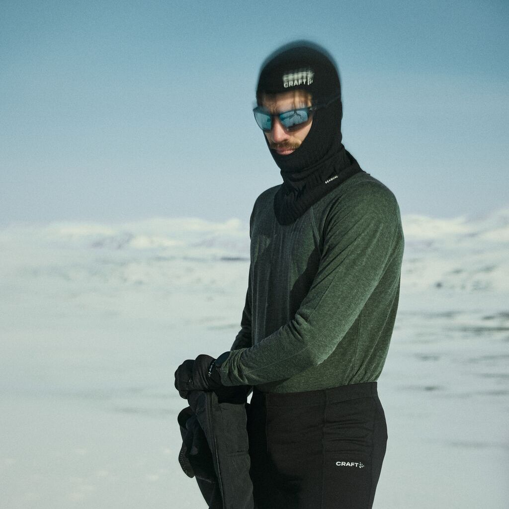 Person i vinterklær, inkludert balaklava og solbriller, stående i et snødekt landskap under klar himmel.